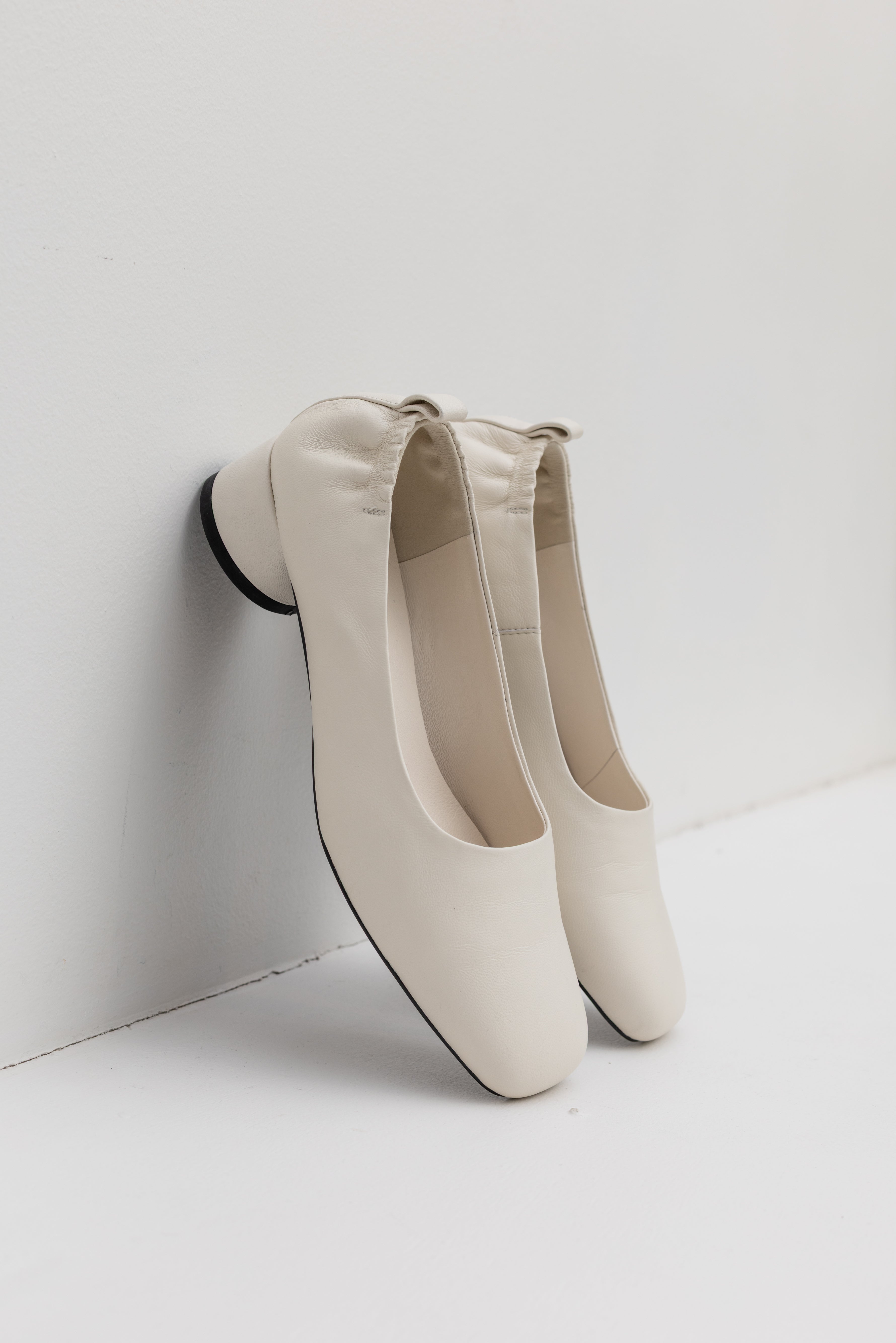A pair of modern and sculptural Amie Rafa label white leather rounded block heel glove pumps with a modern squared off toe and elastic ruched back leaning on a wall at an Alas Eius store. 