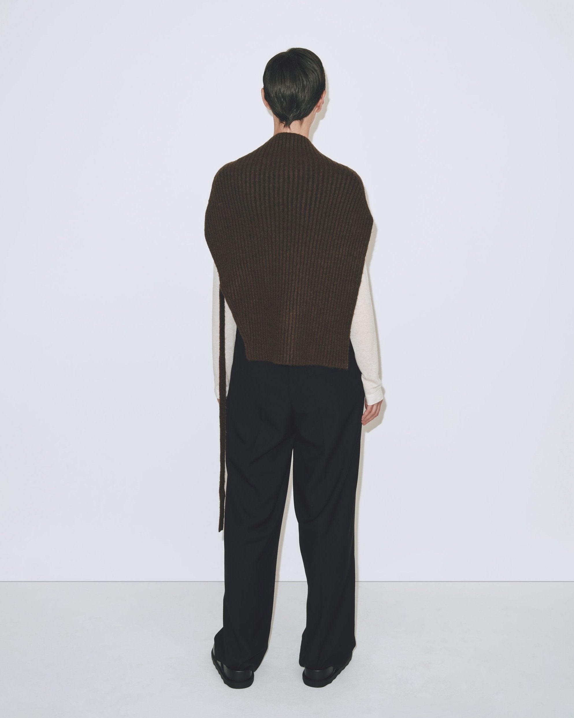 A person wearing an olive brown ribbed knitted Mijeong Park neck warmer with a mock neck neckline and tie waist detail, paired with black trousers at Alas Eius