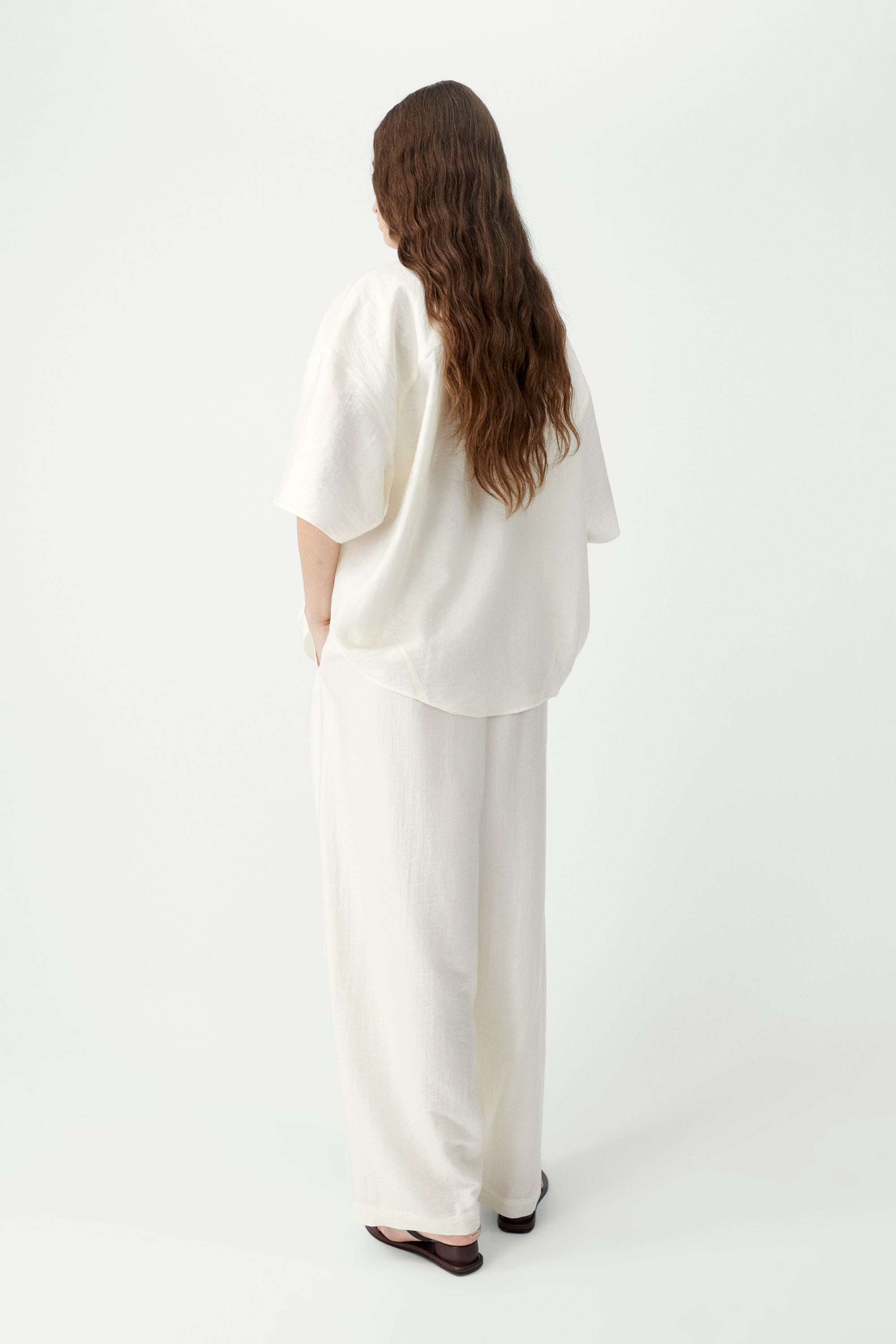 Back view of a woman standing wearing spring fashion in Cordera SS26 Marfil maxi pants designed with a fluid silhouette and a relaxed baggy fit. They feature a drawstring elastic waistband and side pockets at an Alas Eius store. 