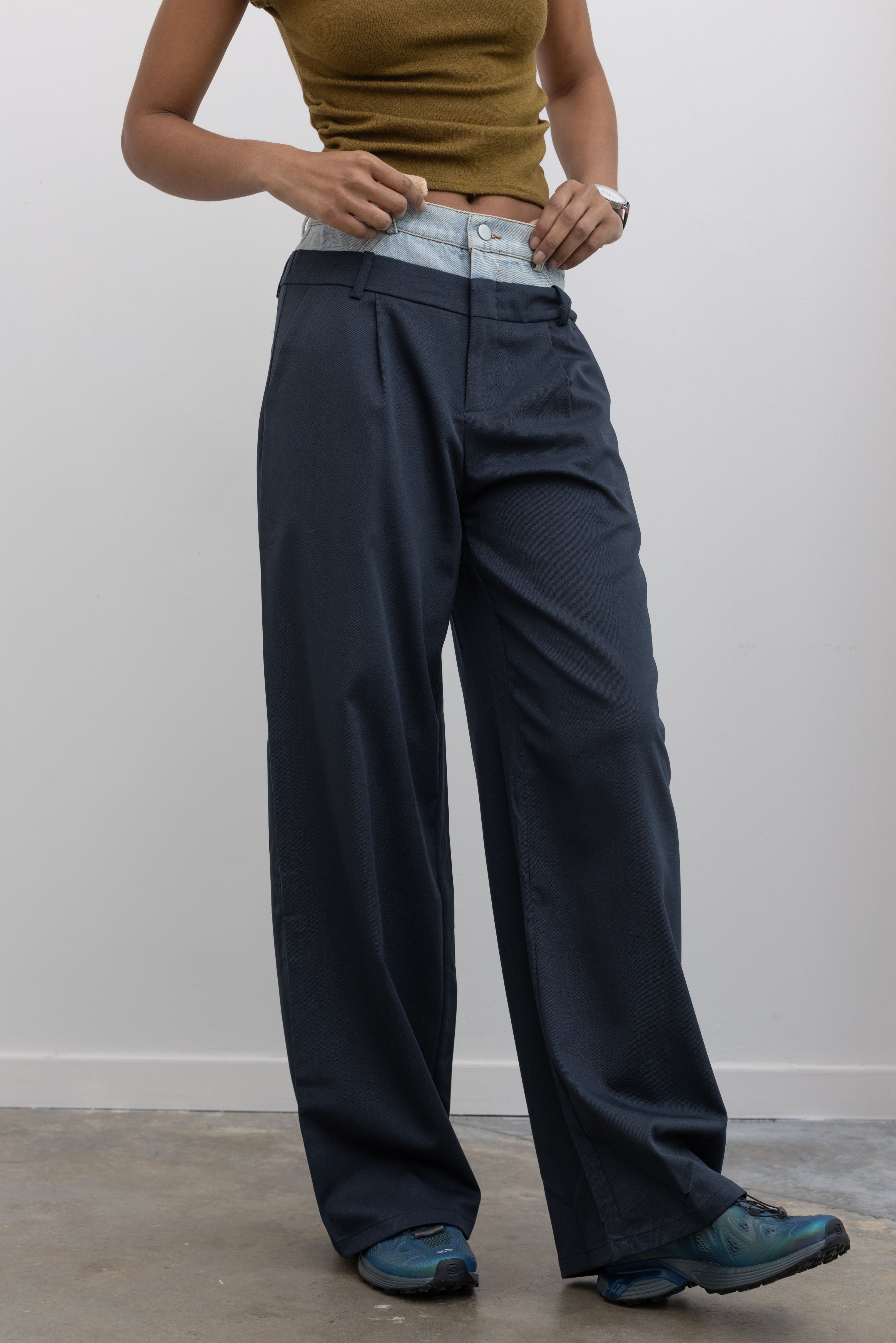 Close-up 3/4 view of a woman wearing spring fashion in Collective Navy Blue pleated trouser featuring a layered look with a light wash denim waistband paired with a short sleeve mock neck top and Salomon sneakers at Alas Eius. 