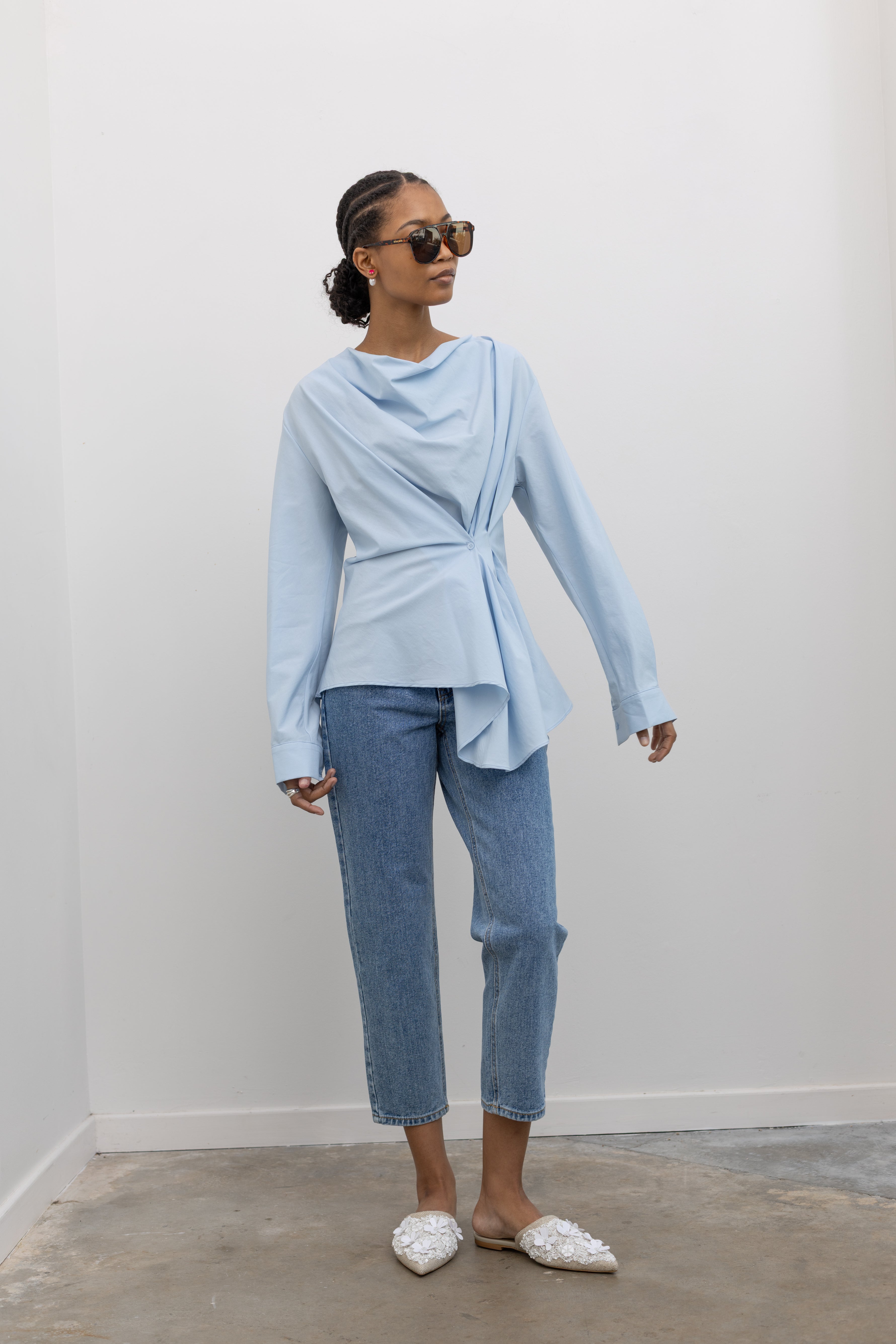 Woman wearing chic and architectural spring fashion in a Collective label sky blue cotton long sleeve top featuring a fitted natural waistline with a side draped motif creating an asymmetrical hem and cowl neckline paired with light wash denim jeans at Alas Eius. 