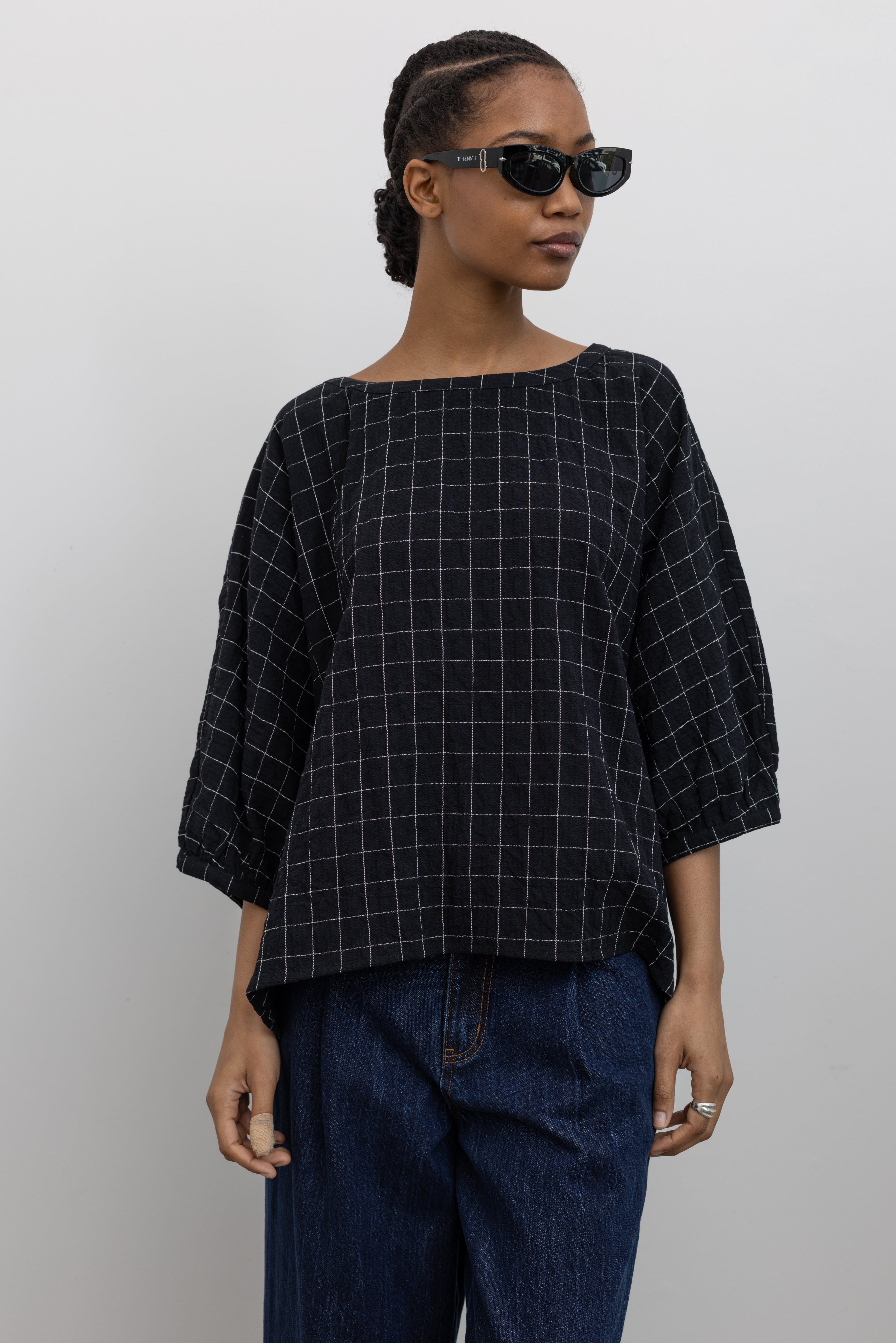 Product view of a woman standing wearing a lightweight checkered cotton gauze oversized top with dropped shoulder seams and 3/4 length sleeves by Collective and paired with dark wash denim jeans at Alas Eius.
