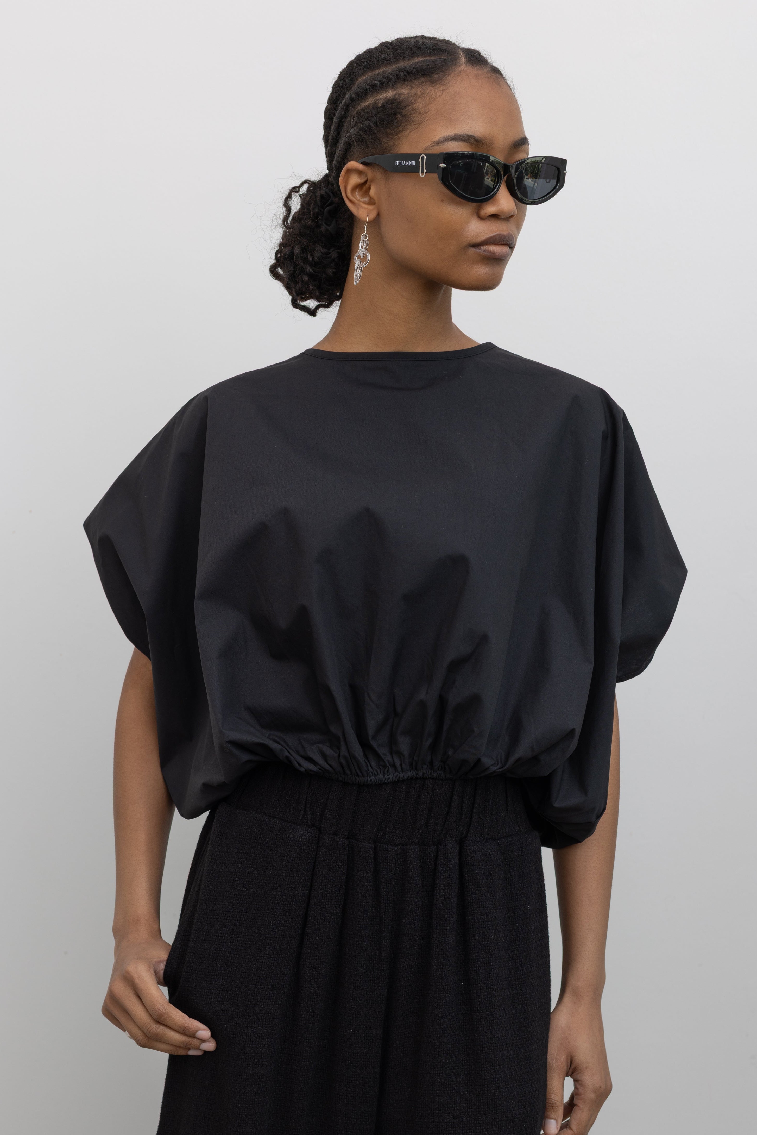 Close-up front view of a woman wearing Spring fashion in a sculptural  Collective Black Cotton billow cinched top featuring draped rounded sleeves, an elasticized waistband for a fitted torso and ribbon tie at the back neck at Alas Eius. 