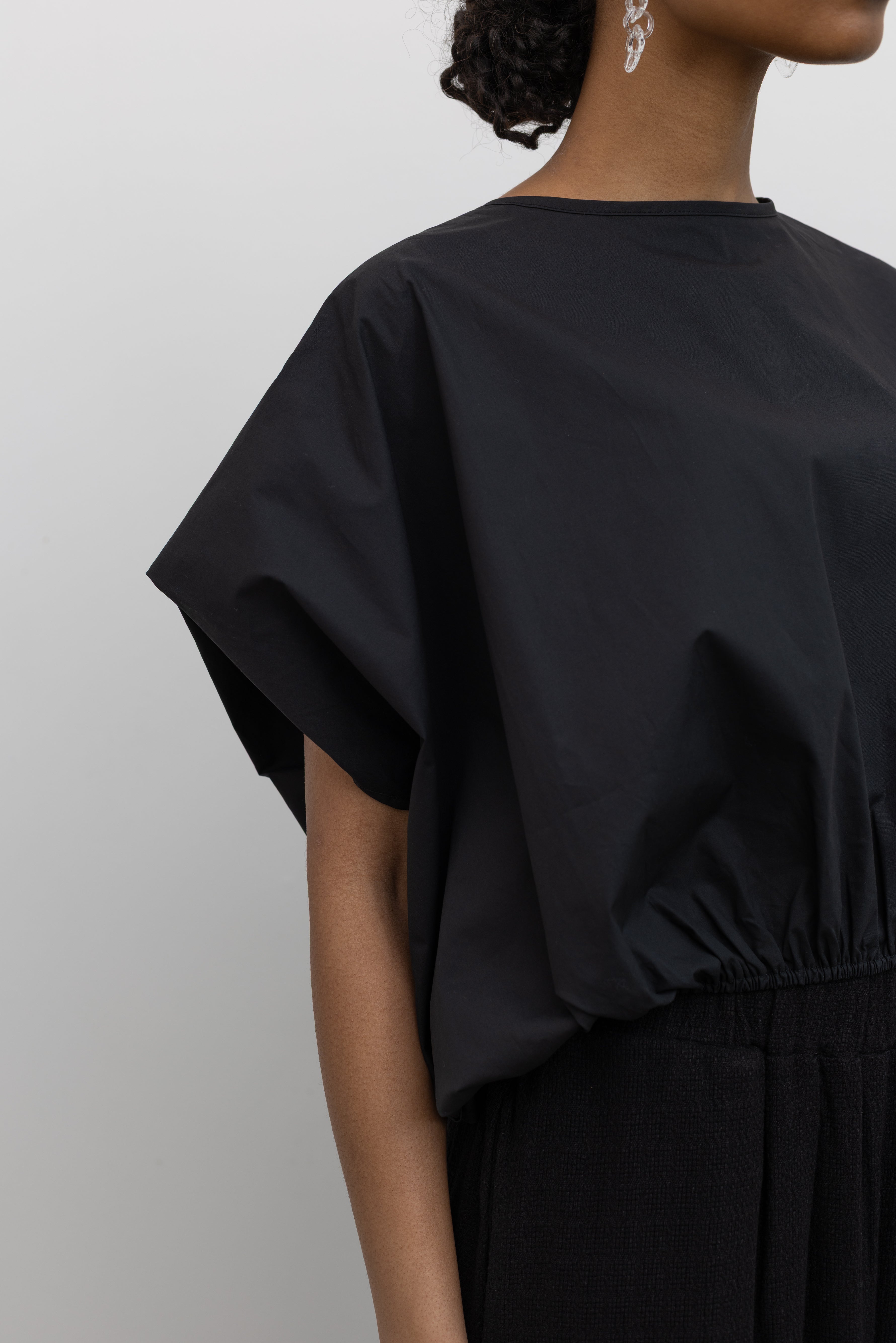 Close-up side view of a woman wearing Spring fashion in a sculptural  Collective Black Cotton billow cinched top featuring draped rounded sleeves, an elasticized waistband for a fitted torso and ribbon tie at the back neck at Alas Eius. 