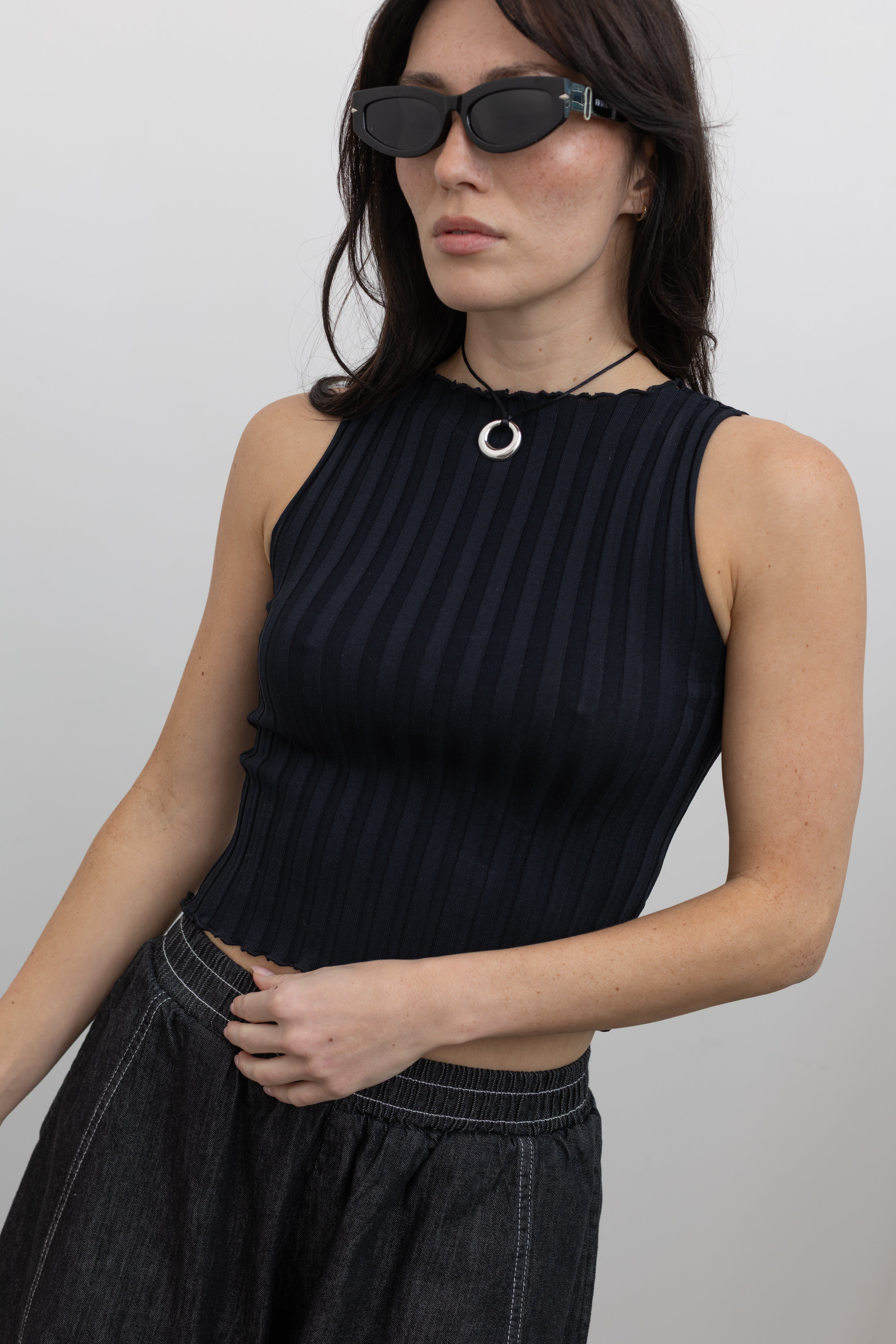Close-up front view of a woman wearing spring capsule wardrobe essential in a Collective brand black fitted ribbed knit lettuce hem sleeveless top Alas Eius Label black cotton barrel leg pull on pants at an Alas Eius store.