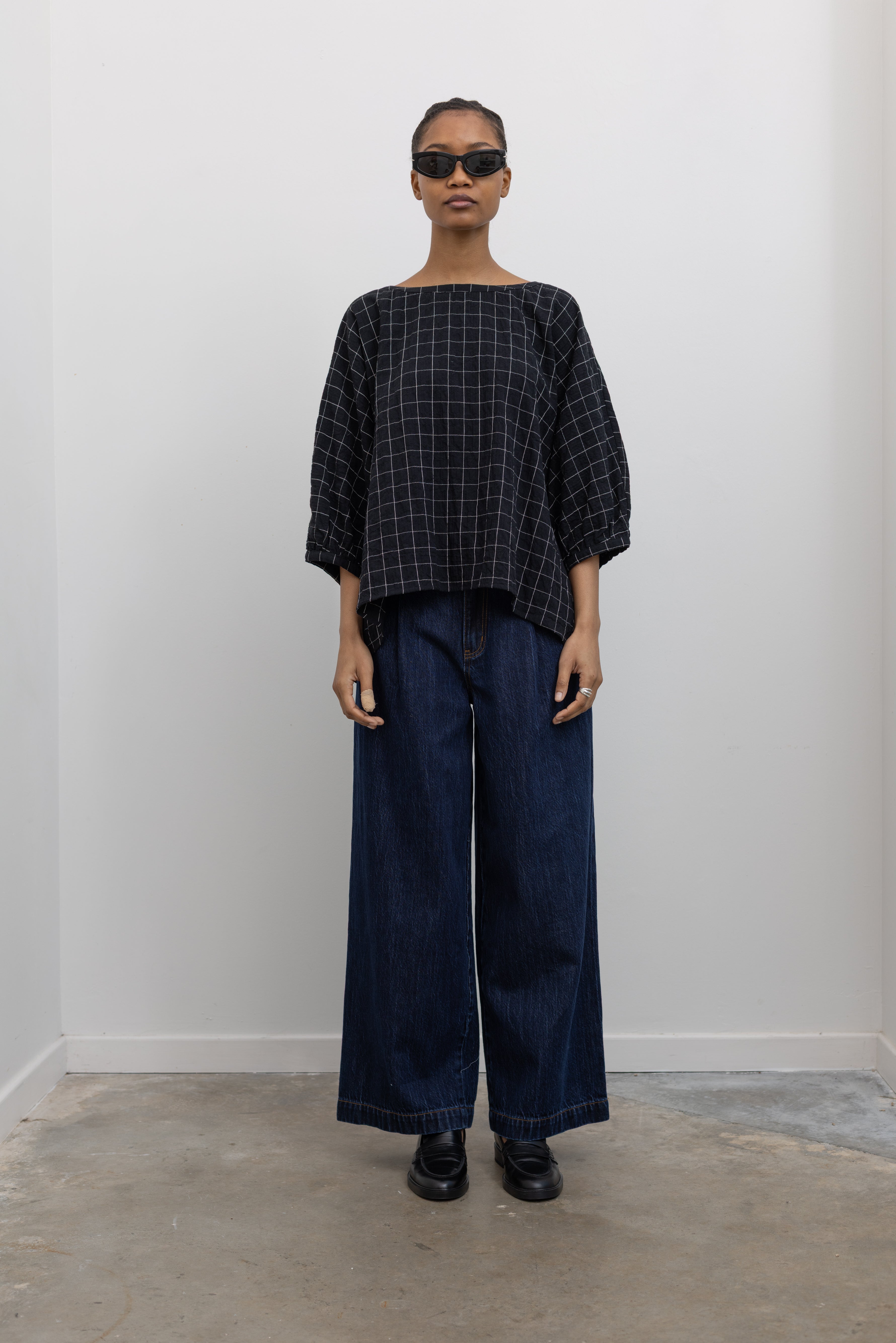 Full front view of a woman standing wearing a lightweight checkered cotton gauze oversized top with dropped shoulder seams and 3/4 length sleeves by Collective and paired with dark wash denim jeans at Alas Eius.