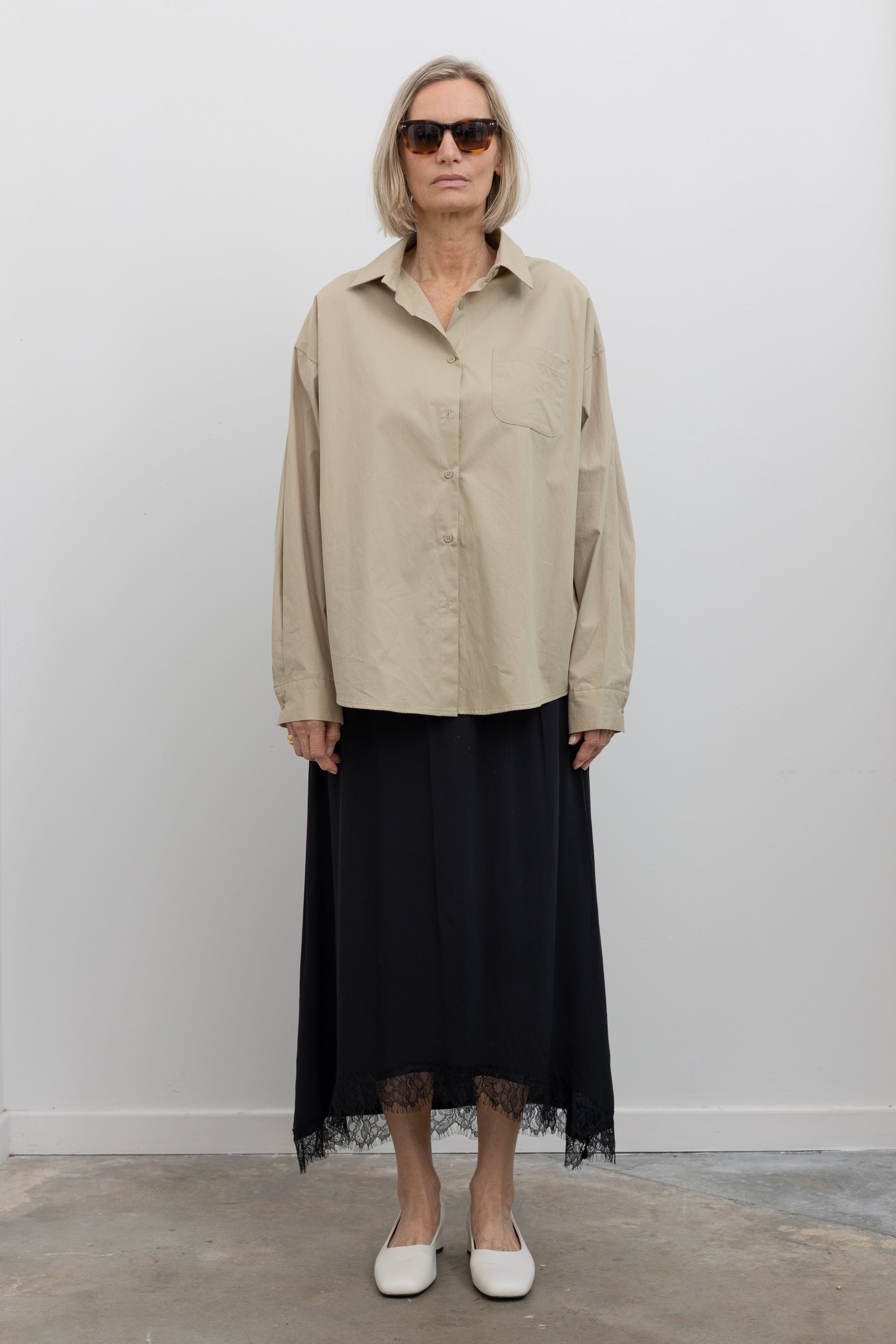 Woman standing wearing a Collective Black Lace Hem Maxi Skirt paired with a classic beige button down shirt and white leather block heels at Alas Eius