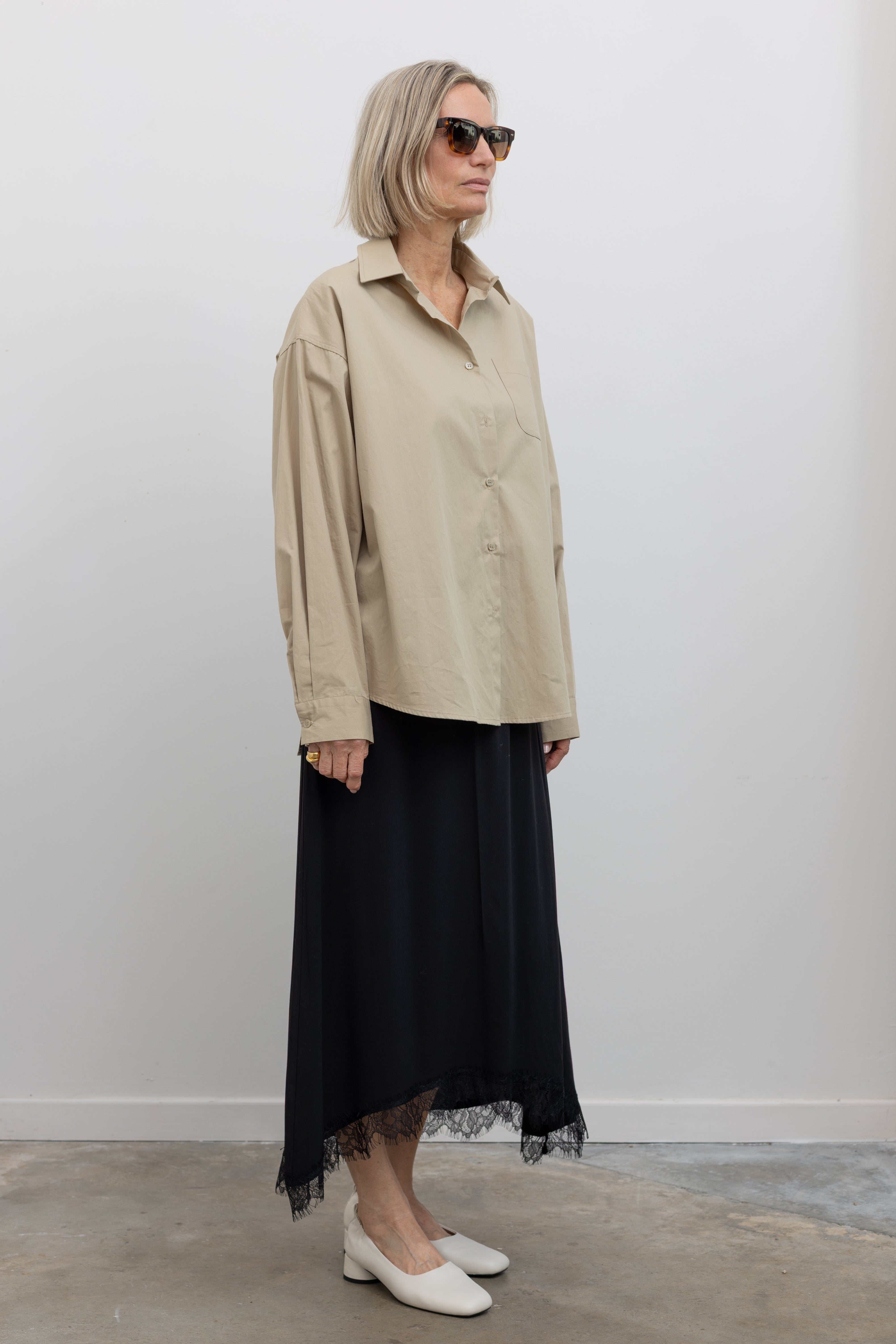 Side view of a woman standing wearing a Collective Black Lace Hem Maxi Skirt paired with a classic beige button down shirt and white leather block heels at Alas Eius