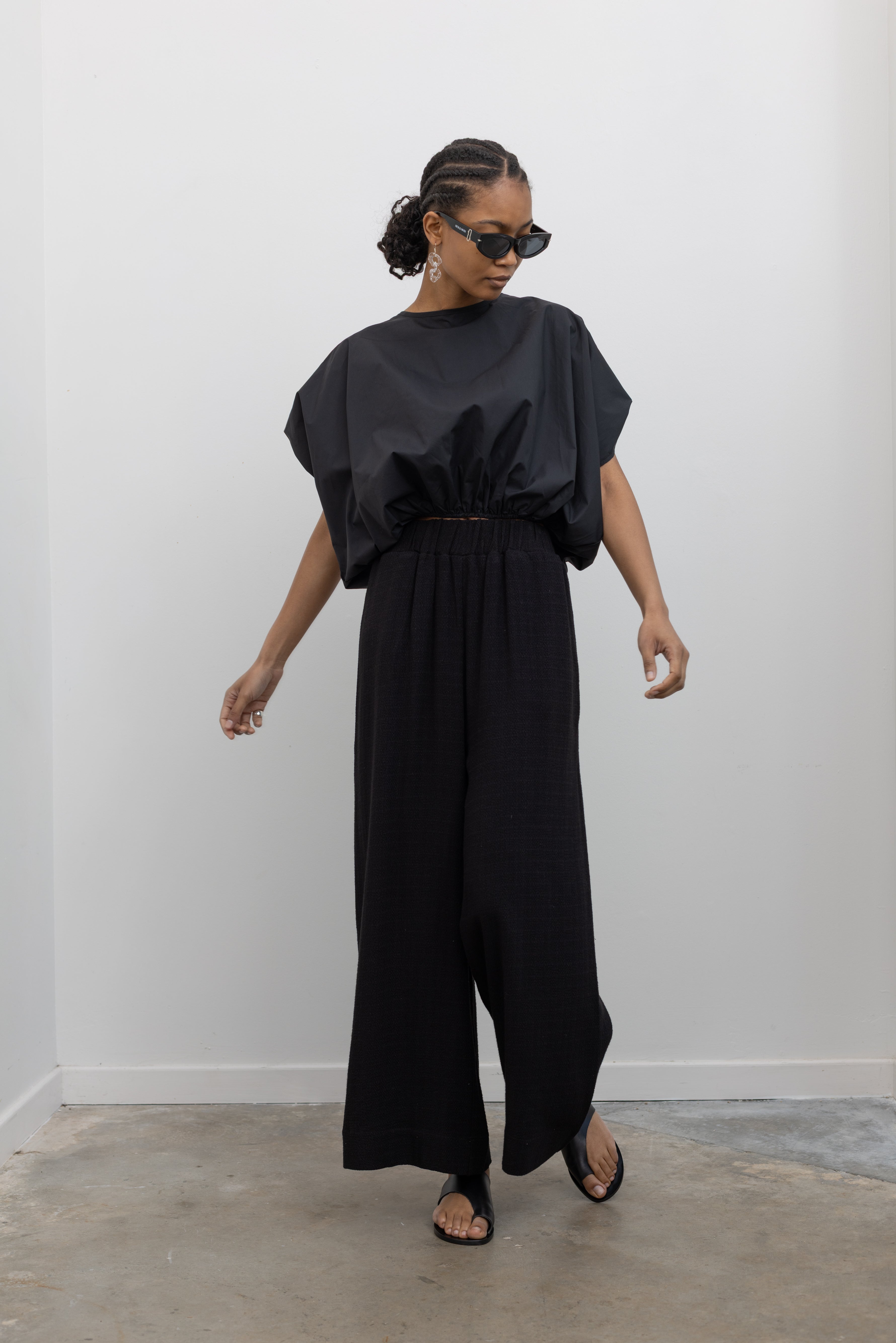 Full length front view of a woman posing wearing contemporary spring fashion in the Collective brand black linen wide leg pull on pants with pockets paired with a black balloon hem top and ATP black leather cutout sandals at an Alas Eius store. 