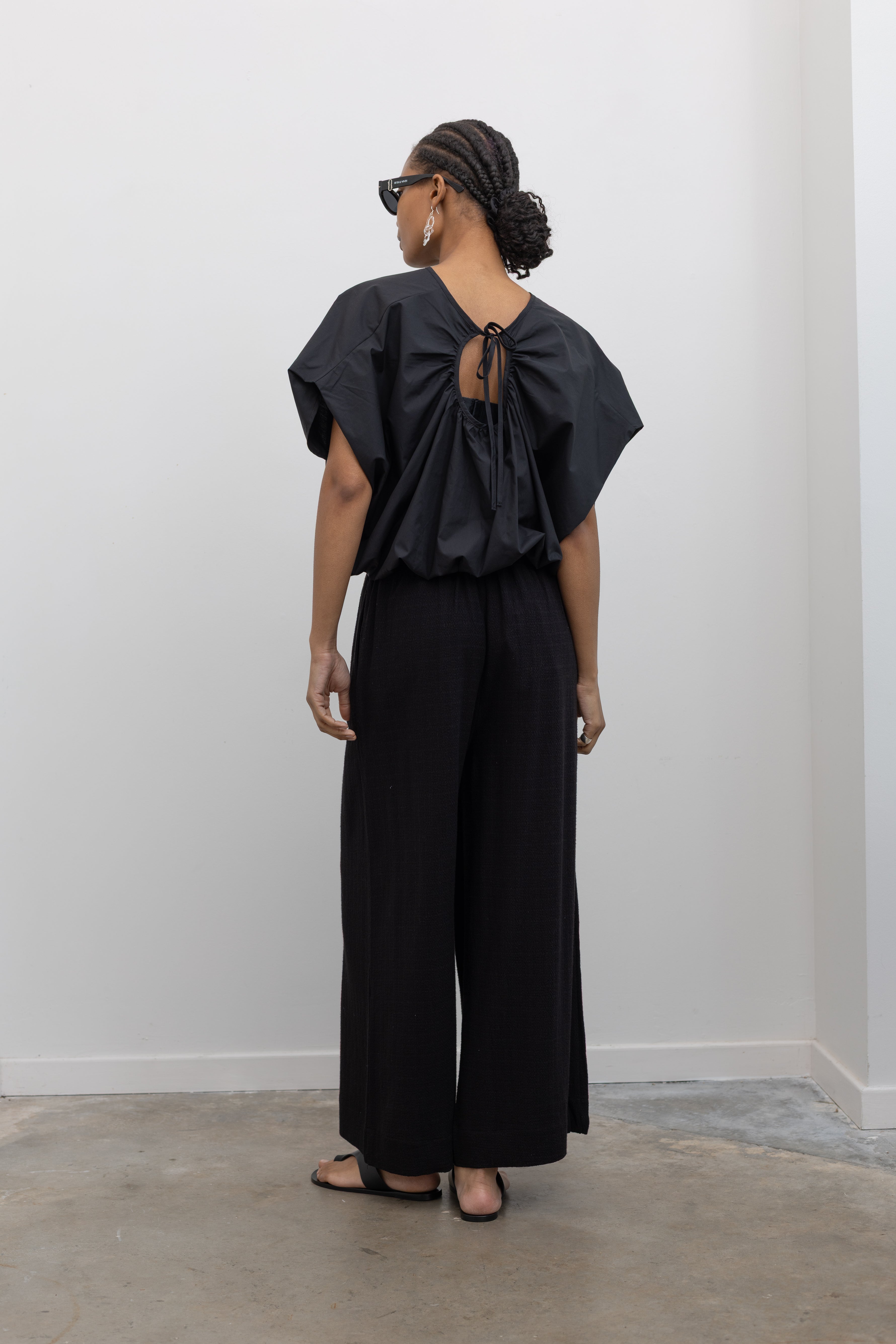 Full length back view of a woman posing wearing contemporary spring fashion in the Collective brand black linen wide leg pull on pants with pockets paired with a black balloon hem top and ATP black leather cutout sandals at an Alas Eius store. 