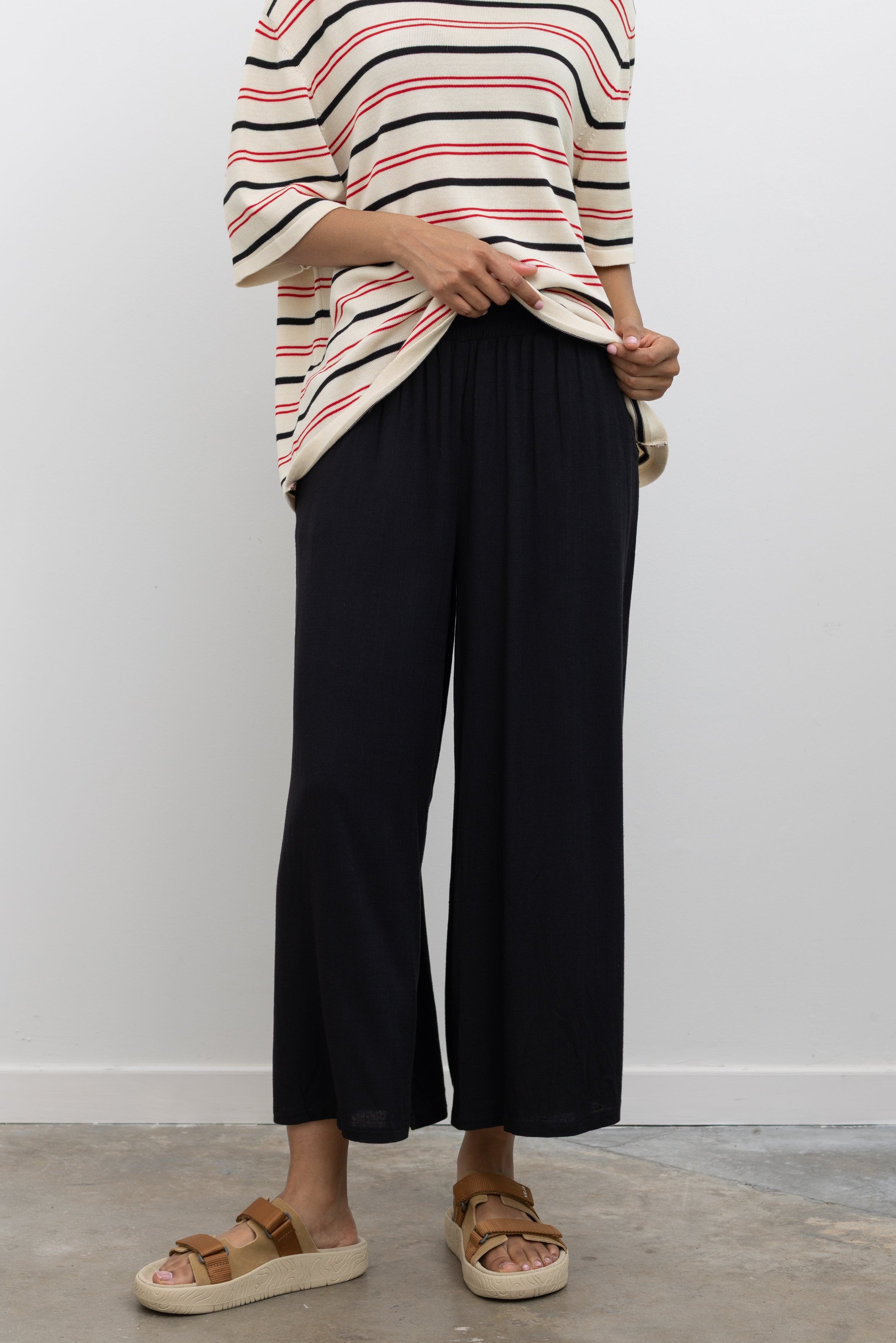 Woman wearing elevated summer weekend fashion in Collective black elastic waistband flowy pull on cropped pants with a Cordera organic cotton striped t-shirt and Veja tan suede slide sandals at Alas Eius. 