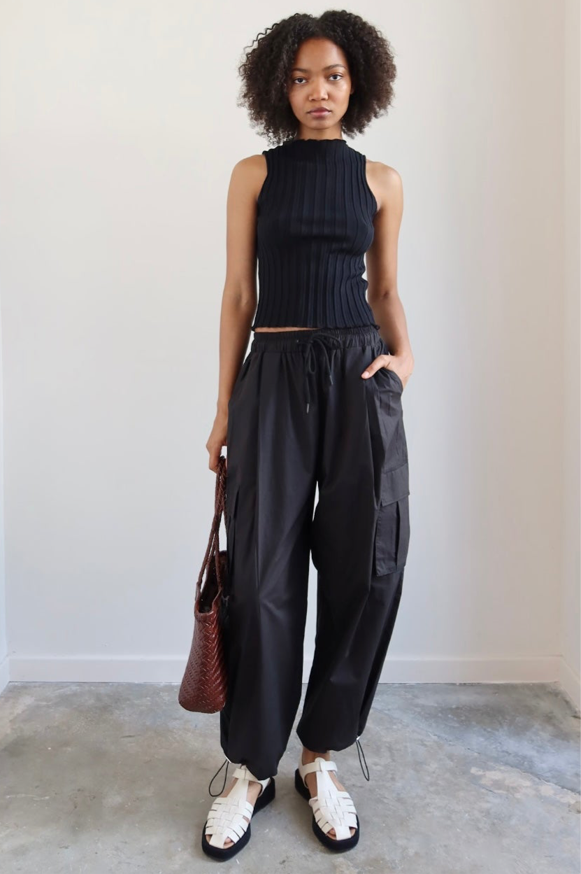 Close-up front view of a woman wearing spring capsule wardrobe essential in a Collective brand black fitted ribbed knit lettuce hem sleeveless top with slight mock neck paired with Alas Eius label black cargo pant and Flattered white leather fisherman platform buckle sandals at Alas Eius. 