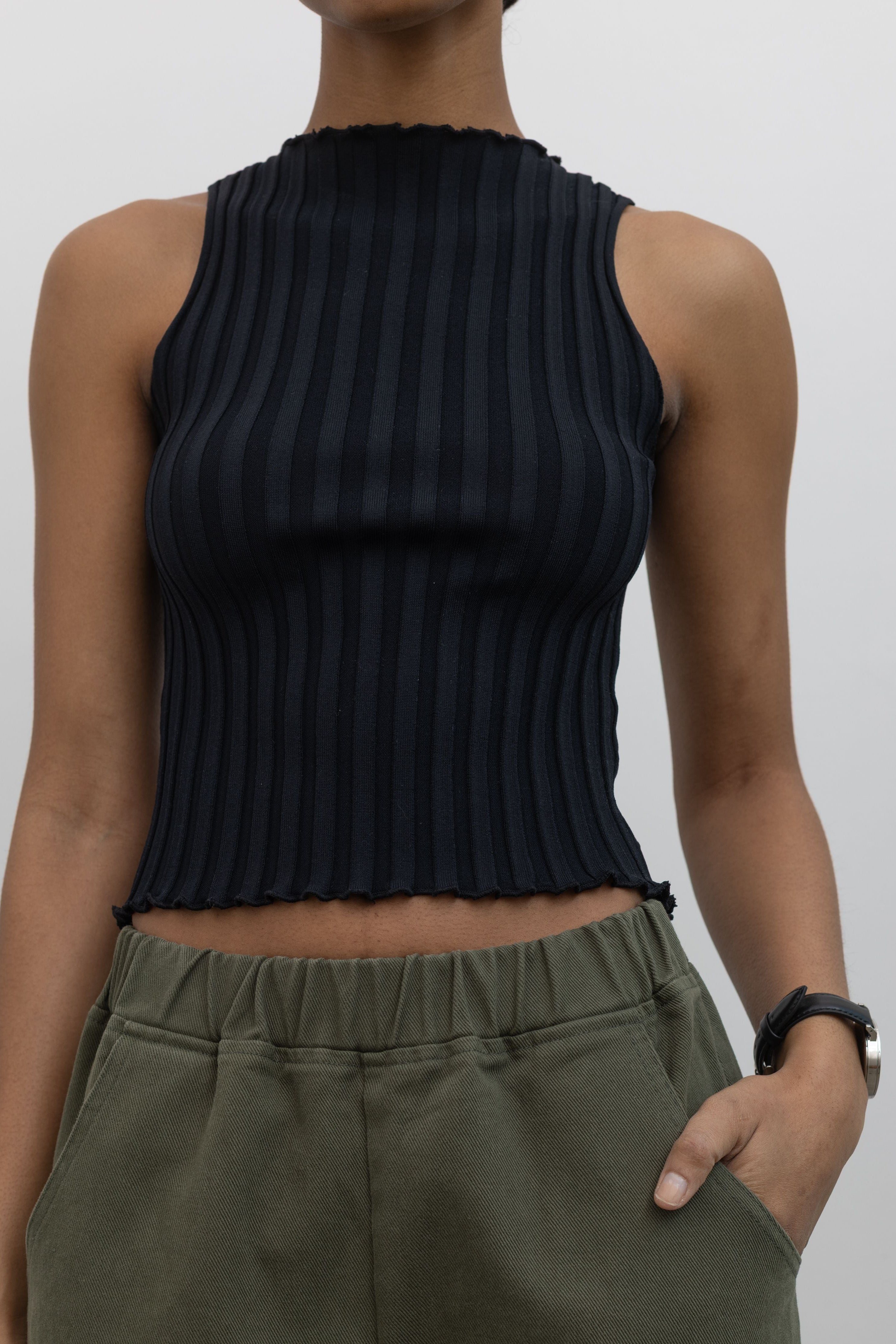 Close-up front view of a woman wearing spring capsule wardrobe essential in a Collective brand black fitted ribbed knit lettuce hem sleeveless top with slight mock neck paired with Le Bon Shoppe olive green cotton canvas arc shaped pant at Alas Eius. 