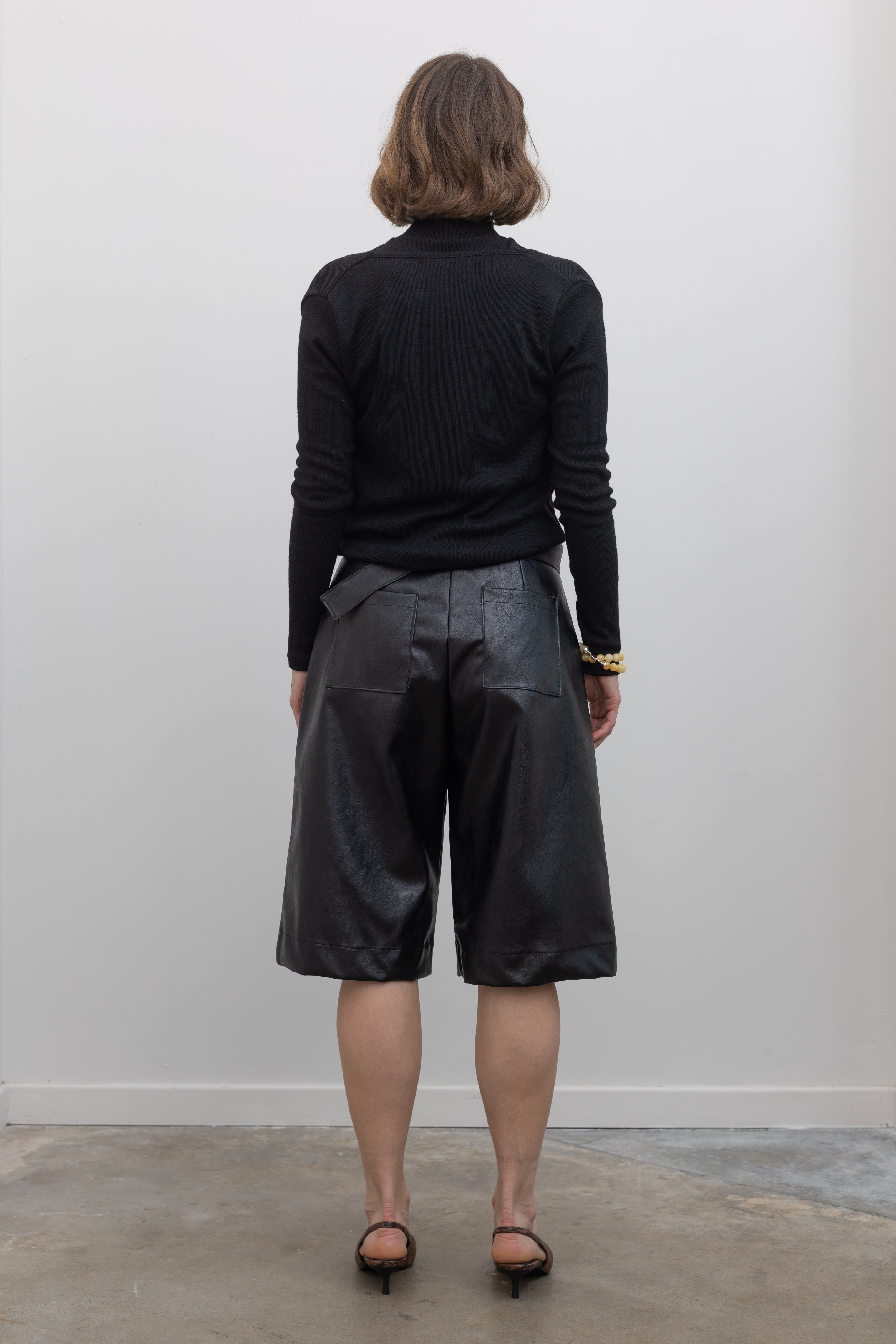 Full length back view of a woman wearing Collective label Black Vegan Leather Belted Bermuda Shorts with an Alas Eius label mock neck sweater and slingback kitten heels at Alas Eius.