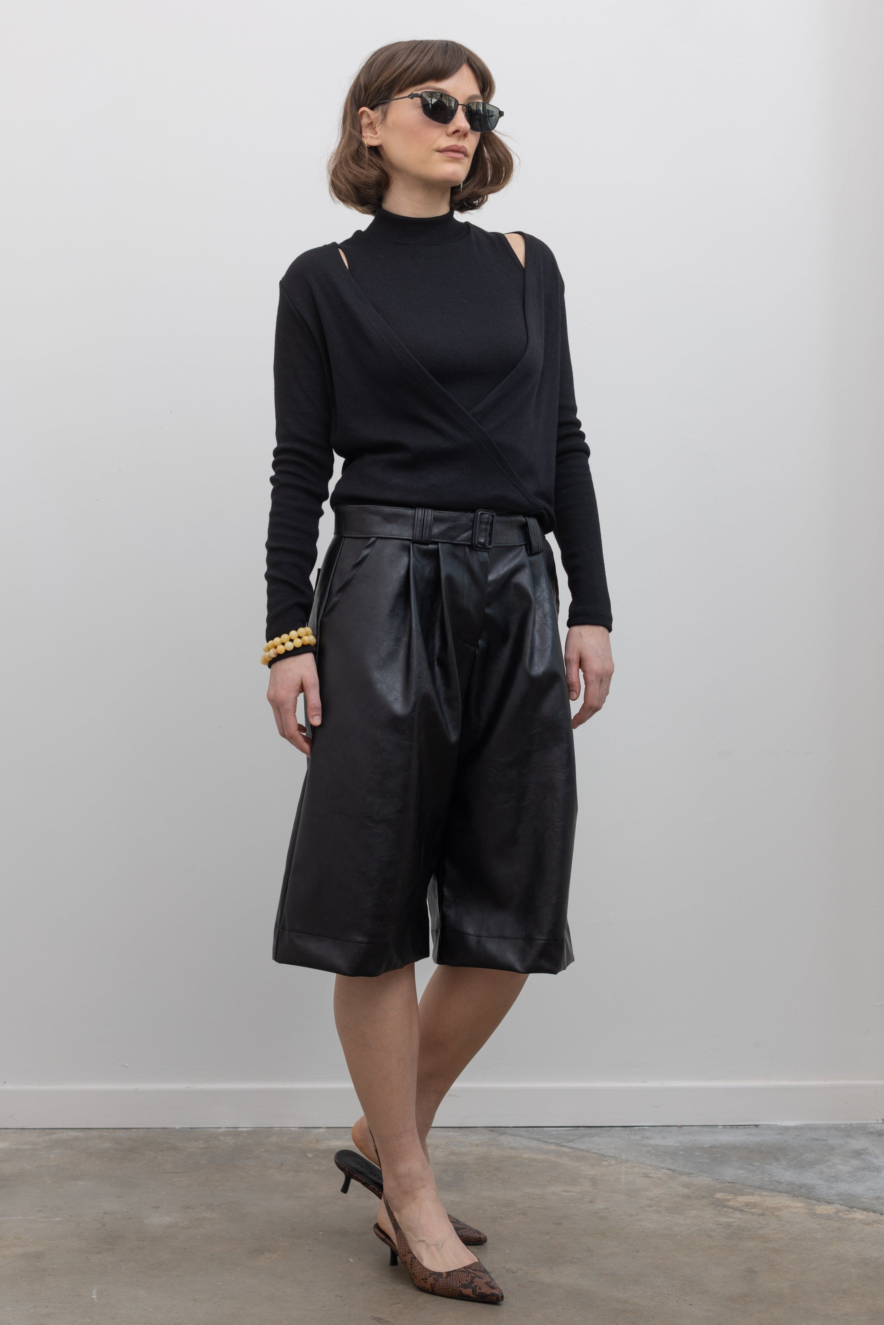 Full length front view of a woman wearing Collective label Black Vegan Leather Belted Bermuda Shorts with an Alas Eius label mock neck sweater and slingback kitten heels at Alas Eius.