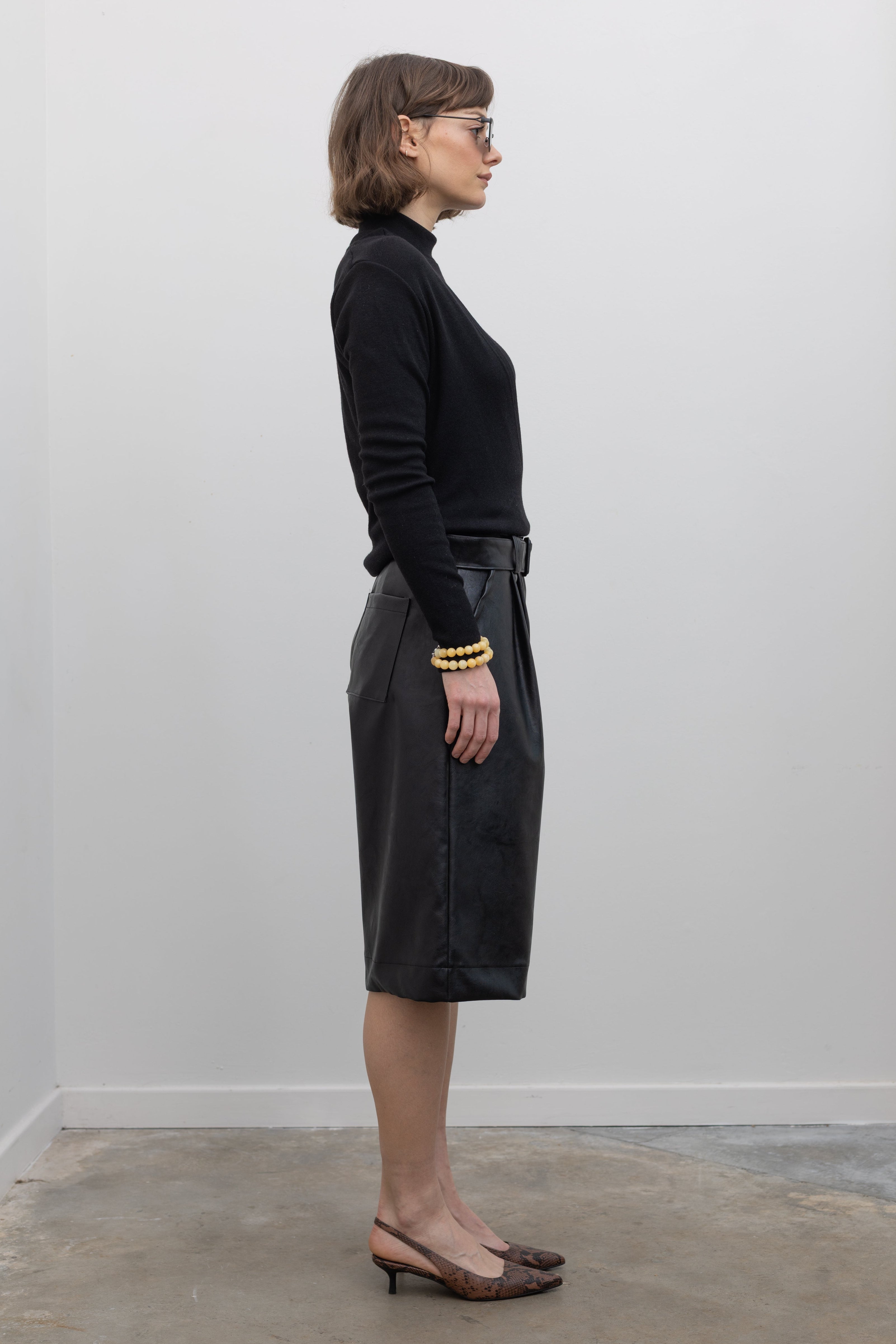 Full length side view of a woman wearing Collective label Black Vegan Leather Belted Bermuda Shorts with an Alas Eius label mock neck sweater and slingback kitten heels at Alas Eius.