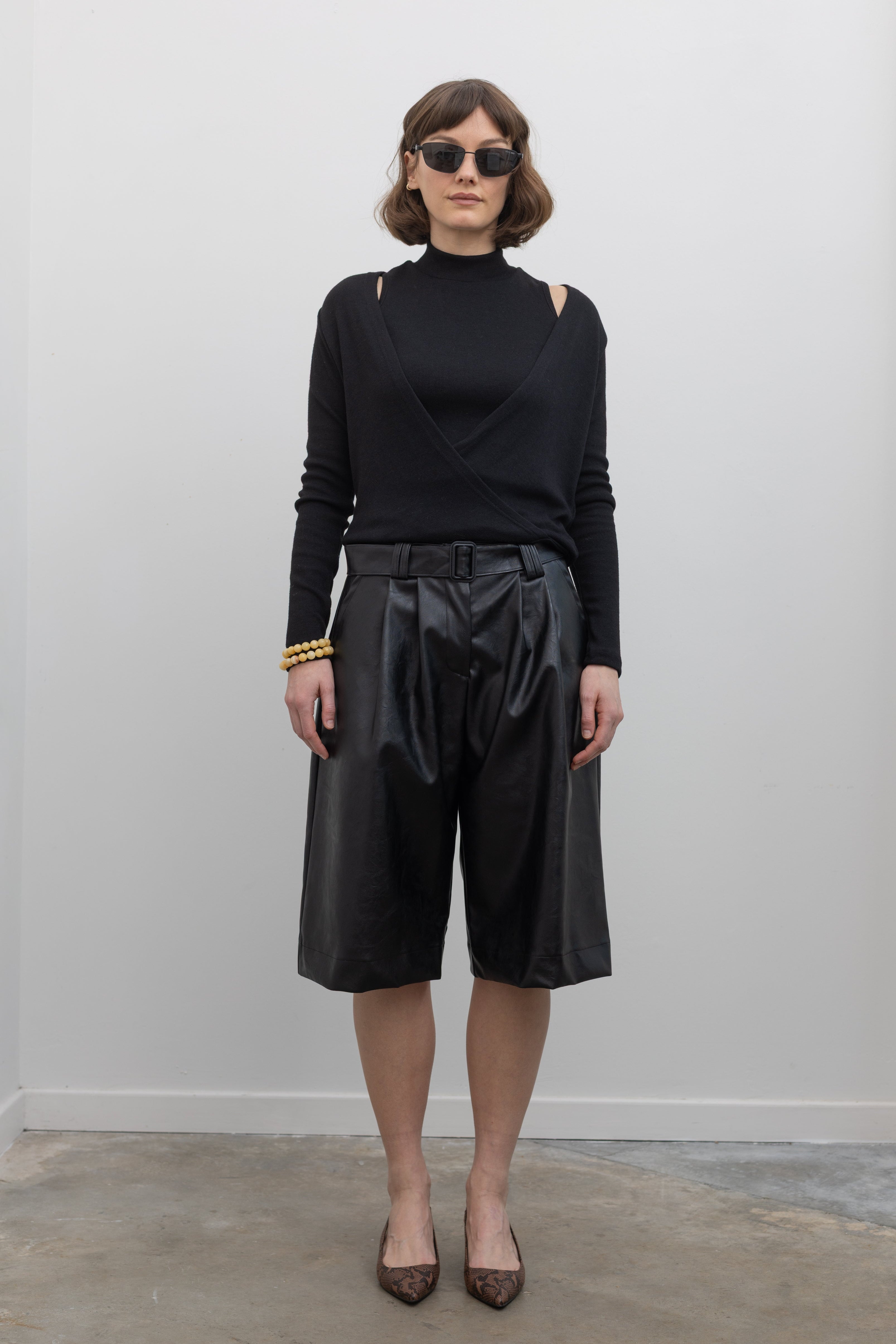 Full length front view of a woman wearing Collective label Black Vegan Leather Belted Bermuda Shorts with an Alas Eius label mock neck sweater and slingback kitten heels at Alas Eius.