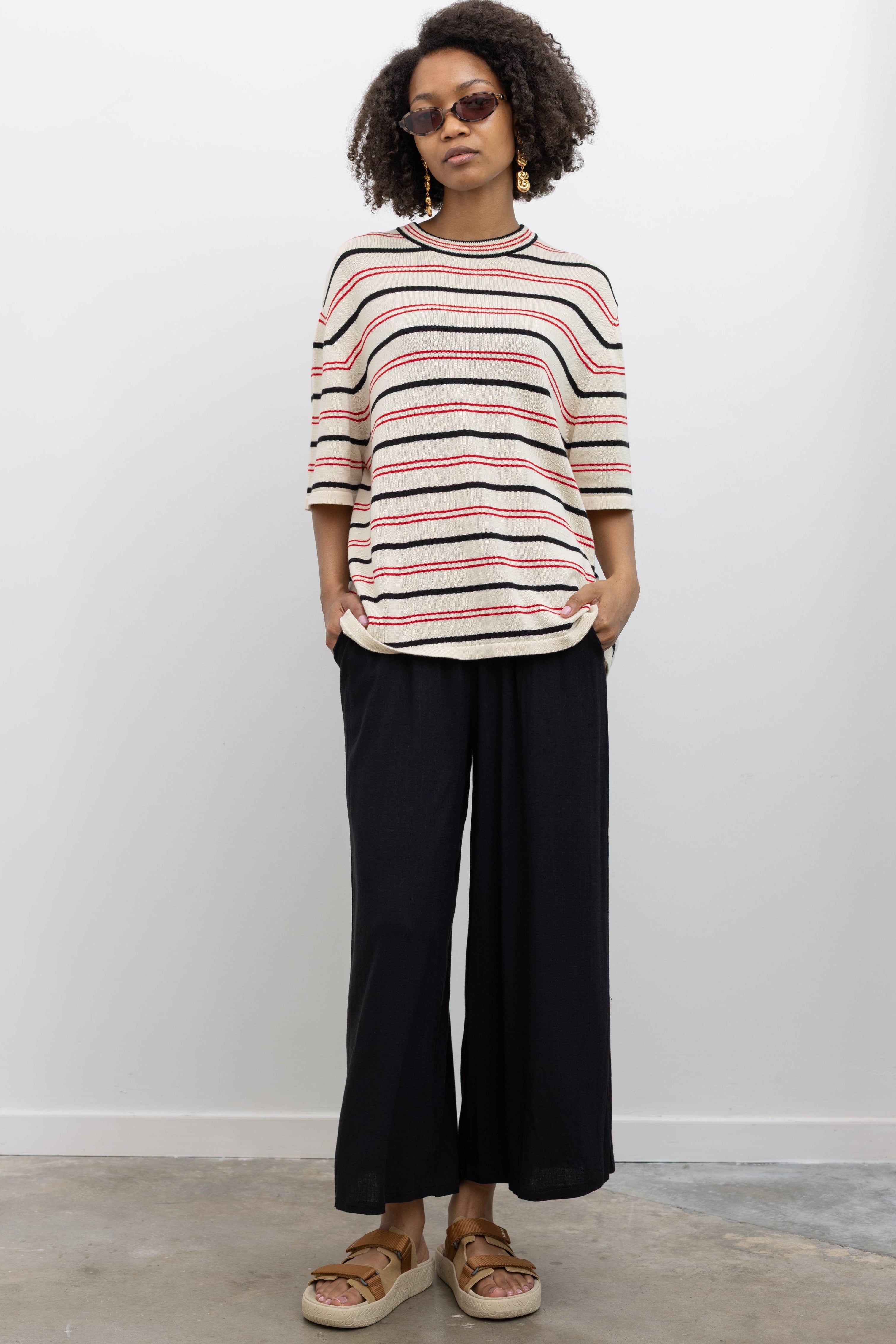 Woman wearing modern minimalist fashion in Collective black elastic waistband flowy pull on cropped pants with a Cordera organic cotton striped t-shirt and Veja tan suede slide sandals at Alas Eius. 