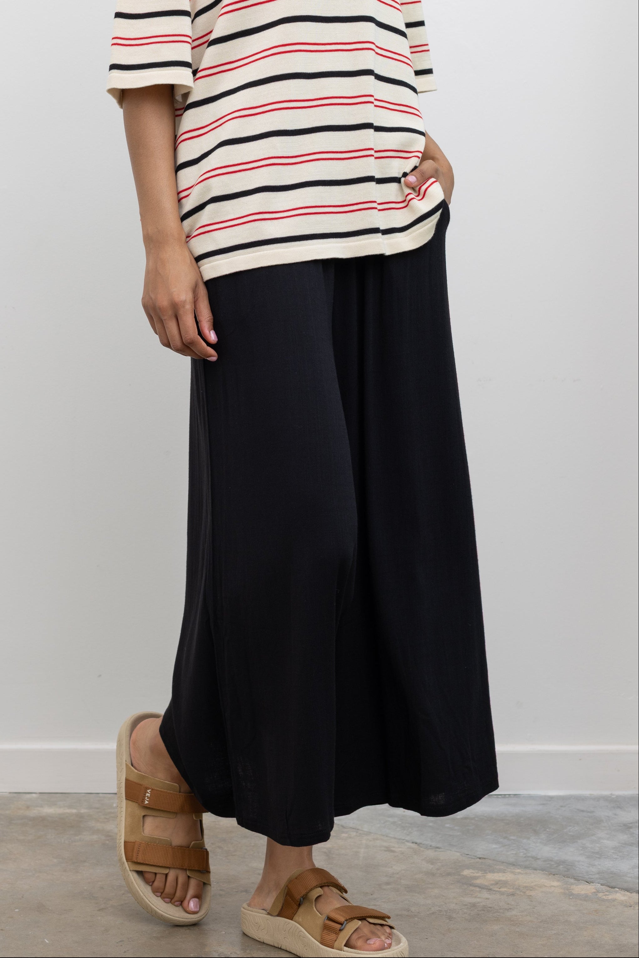 Woman wearing chic summer fashion in Collective black elastic waistband flowy pull on cropped pants with a Cordera organic cotton striped t-shirt and Veja tan suede slide sandals at Alas Eius. 