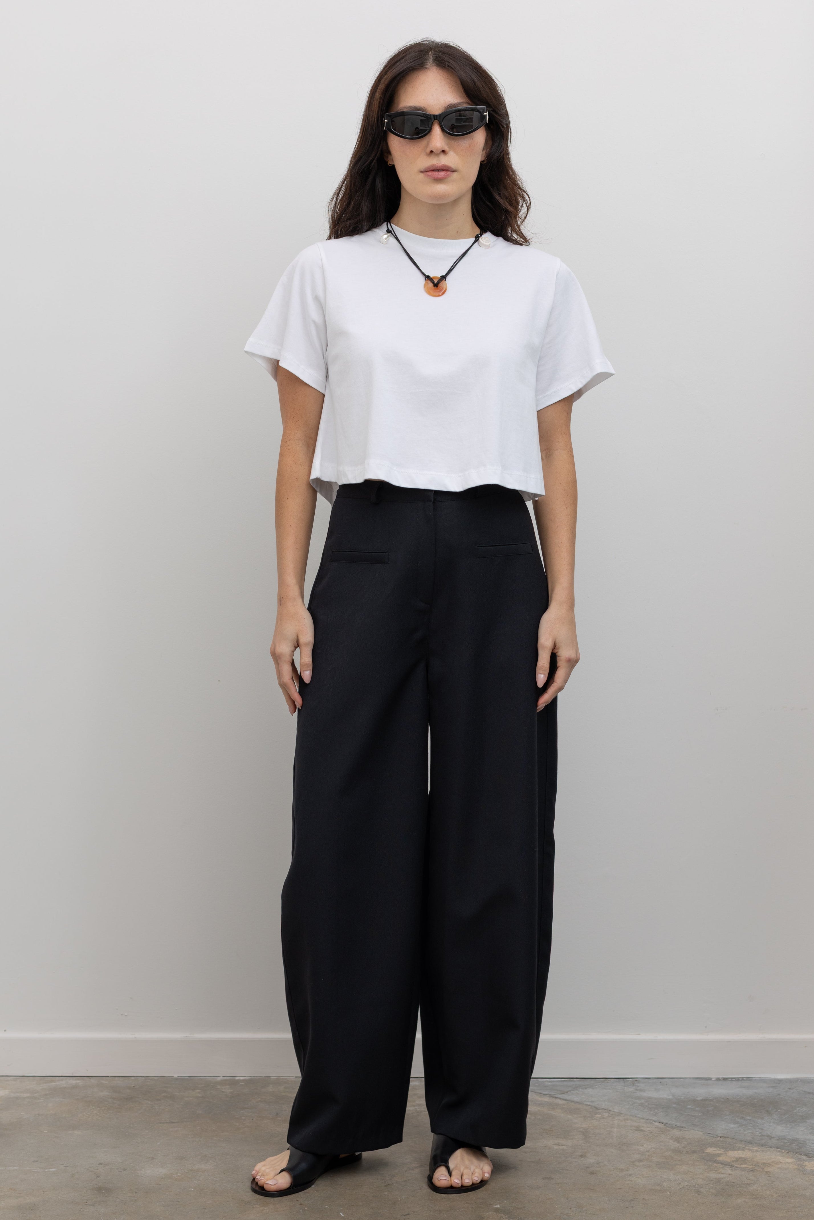 Full length front view of a woman standing wearing chic spring fashion in a Collective black wool carrot high-waisted, wide-leg trouser paired with a white boxy cropped top at Alas Eius. 