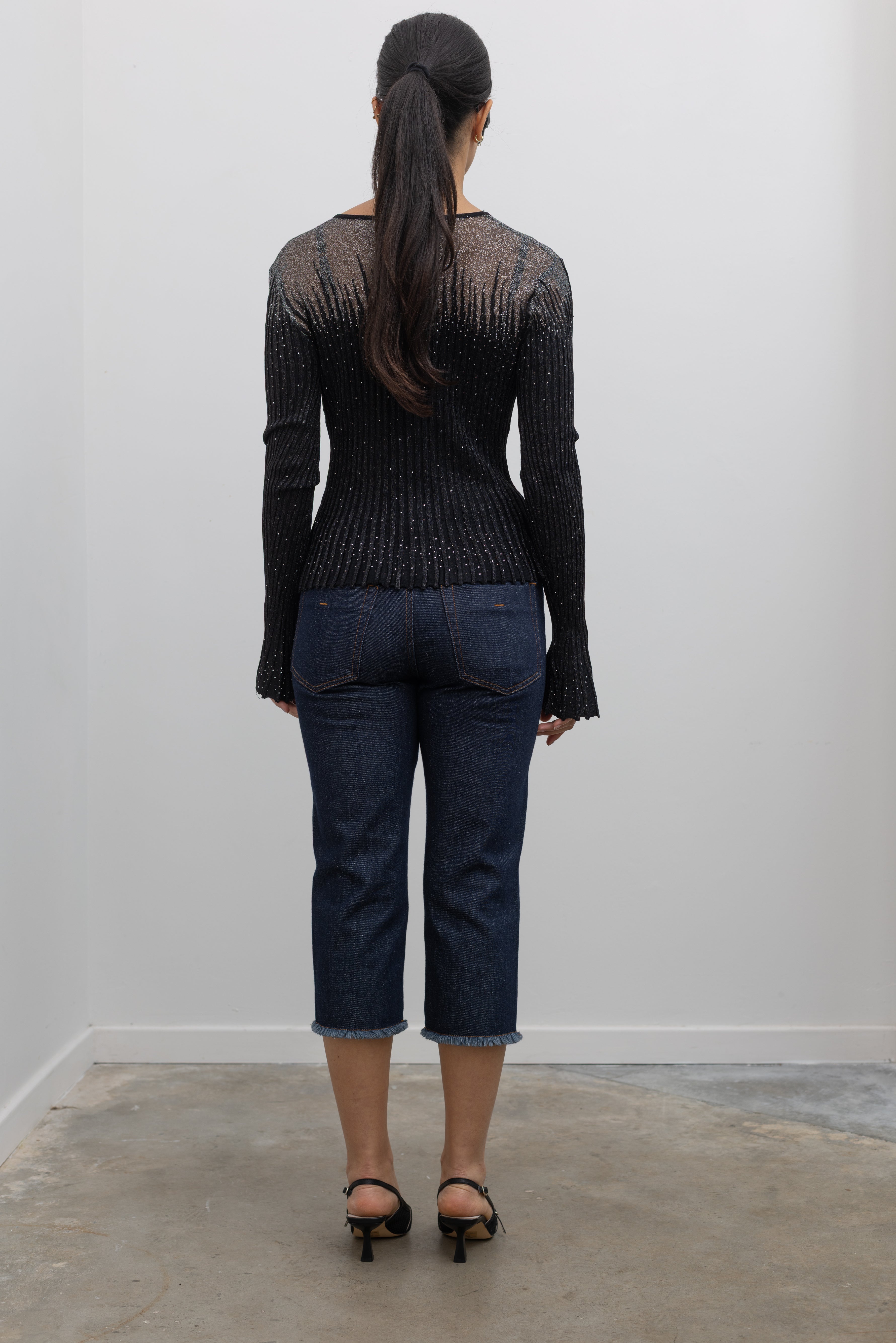 Back view of a woman wearing a Collective Label black top featuring a metallic jeweled motif throughout with long bell sleeves and a faux button down front paired with dark wash denim jeans at an Alas Eius.