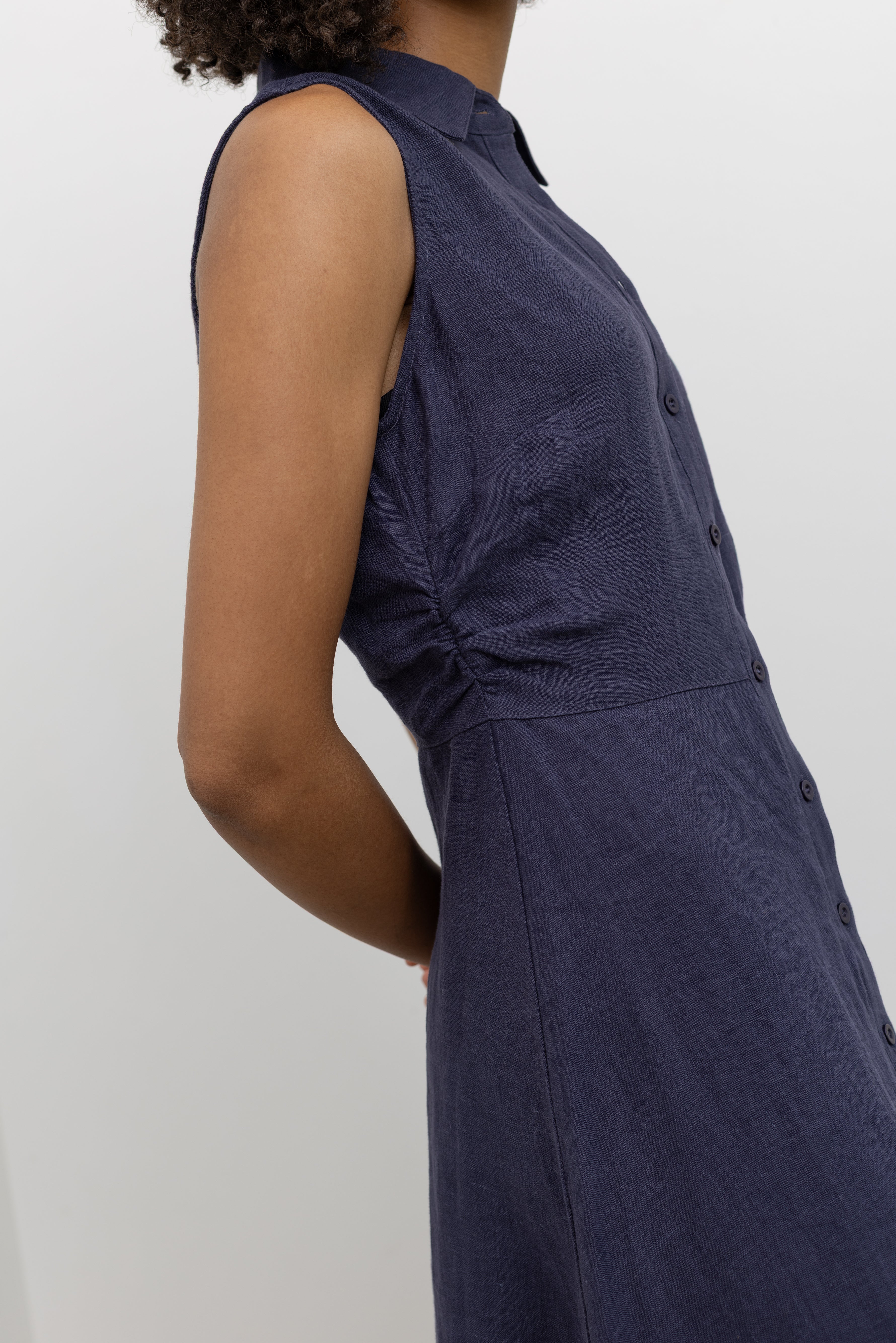 Side view of the Collective navy blue linen sleeveless shirt dress with flattering side ruching details at Alas Eius