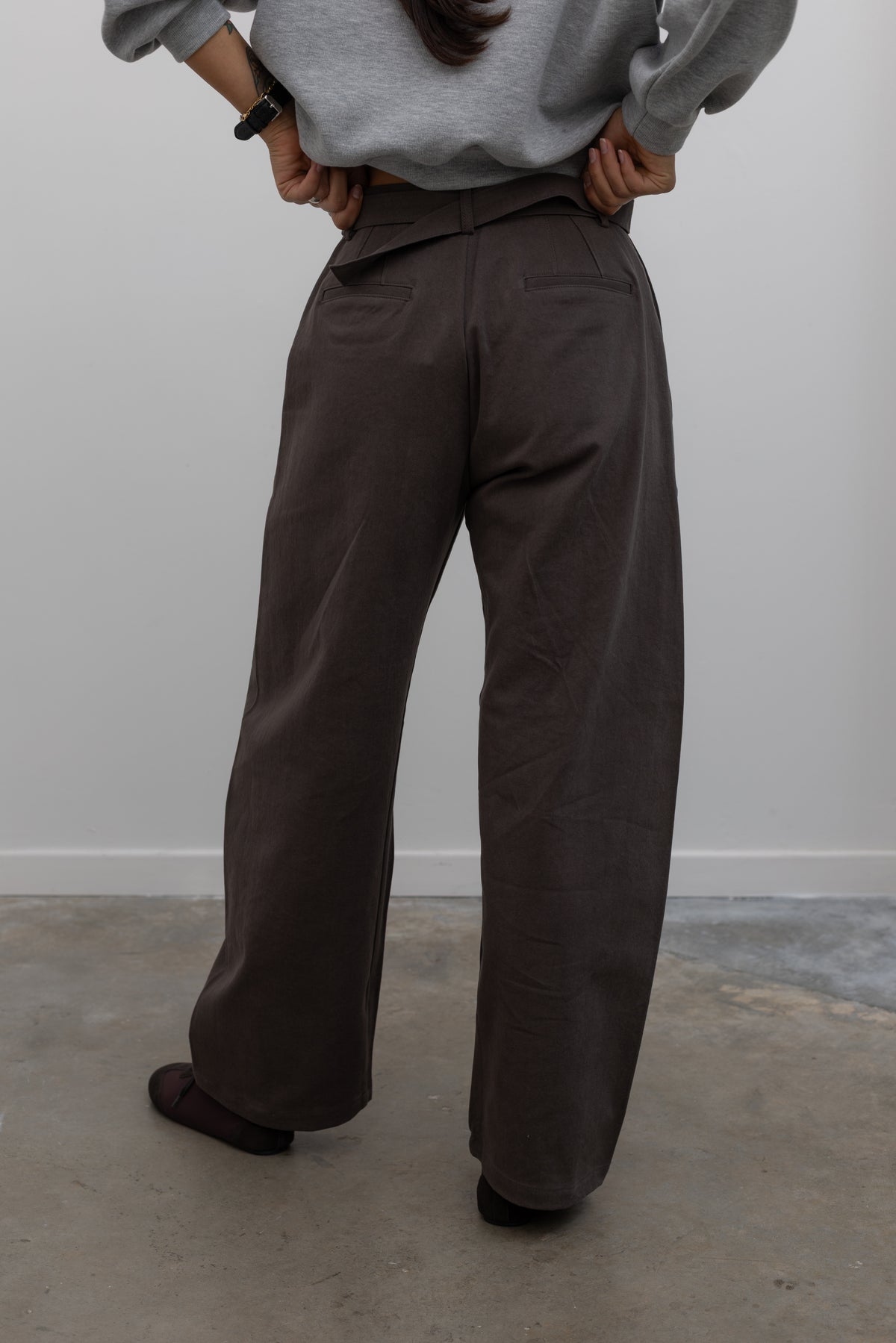 Close-up back view of a woman wearing weekend fashion in a pair of Collective brand brown curved leg belted chino pants with a casual heather gray sweatshirt and brown suede ballet inspired sneakers at an Alas Eius store. 