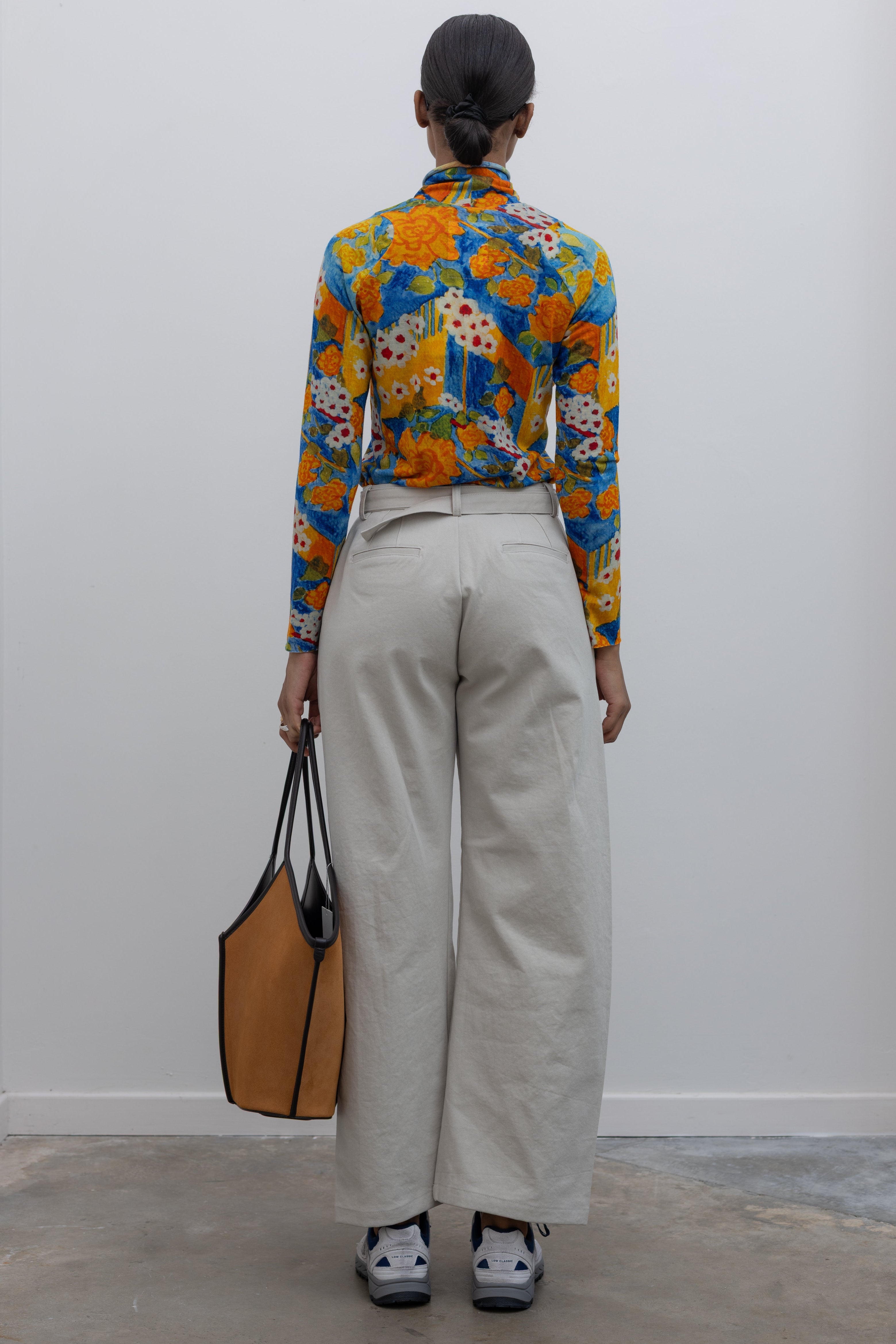Back view of a woman wearing weekend fashion in a pair of Collective brand light gray curved leg belted chino pants with a vibrant orange and blue floral printed long sleeve top paired with white and blue sneakers at Alas Eius.