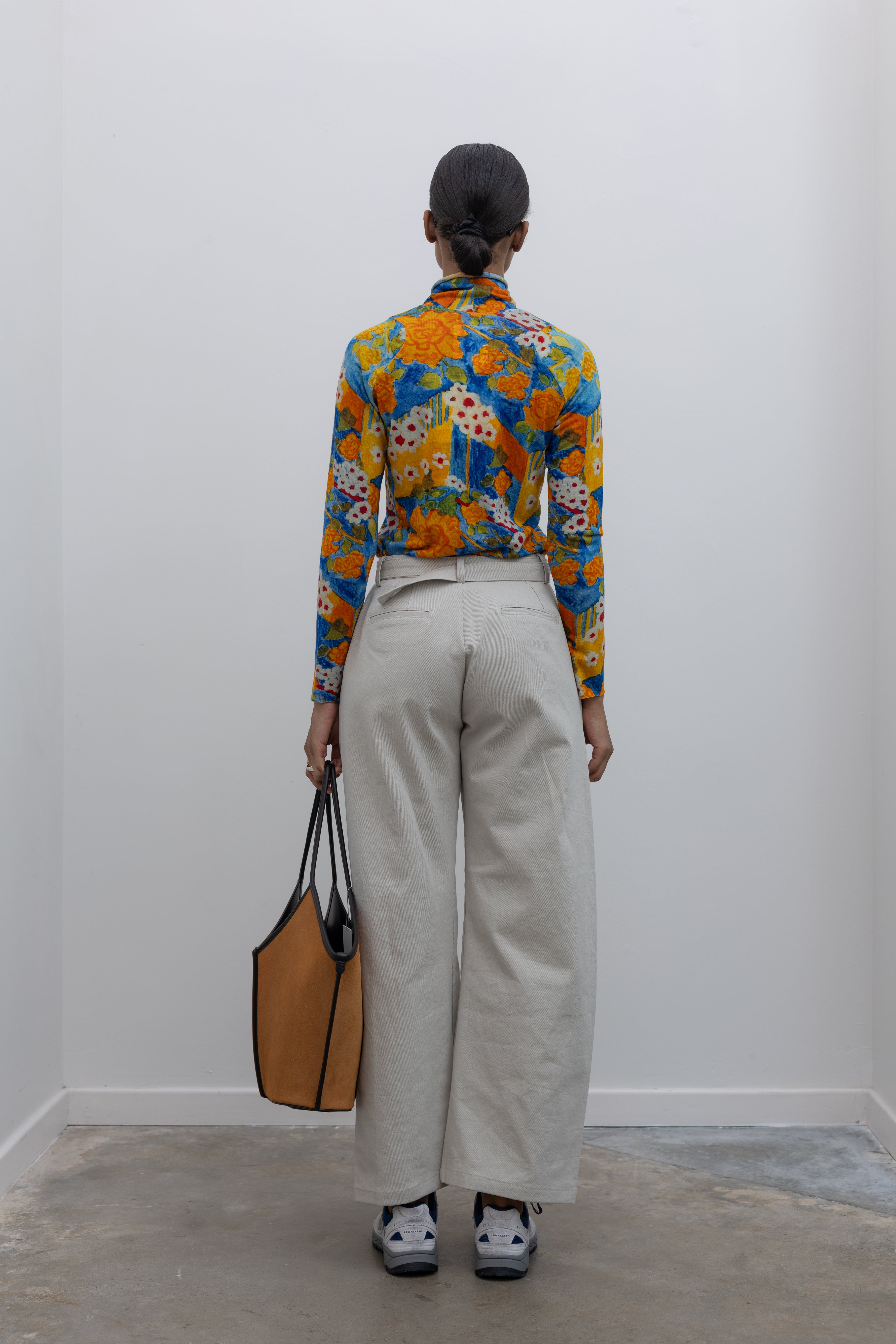 Back view of a woman wearing weekend fashion in a pair of Collective brand light gray curved leg belted chino pants with a vibrant orange and blue floral printed long sleeve top paired with white and blue sneakers at Alas Eius.