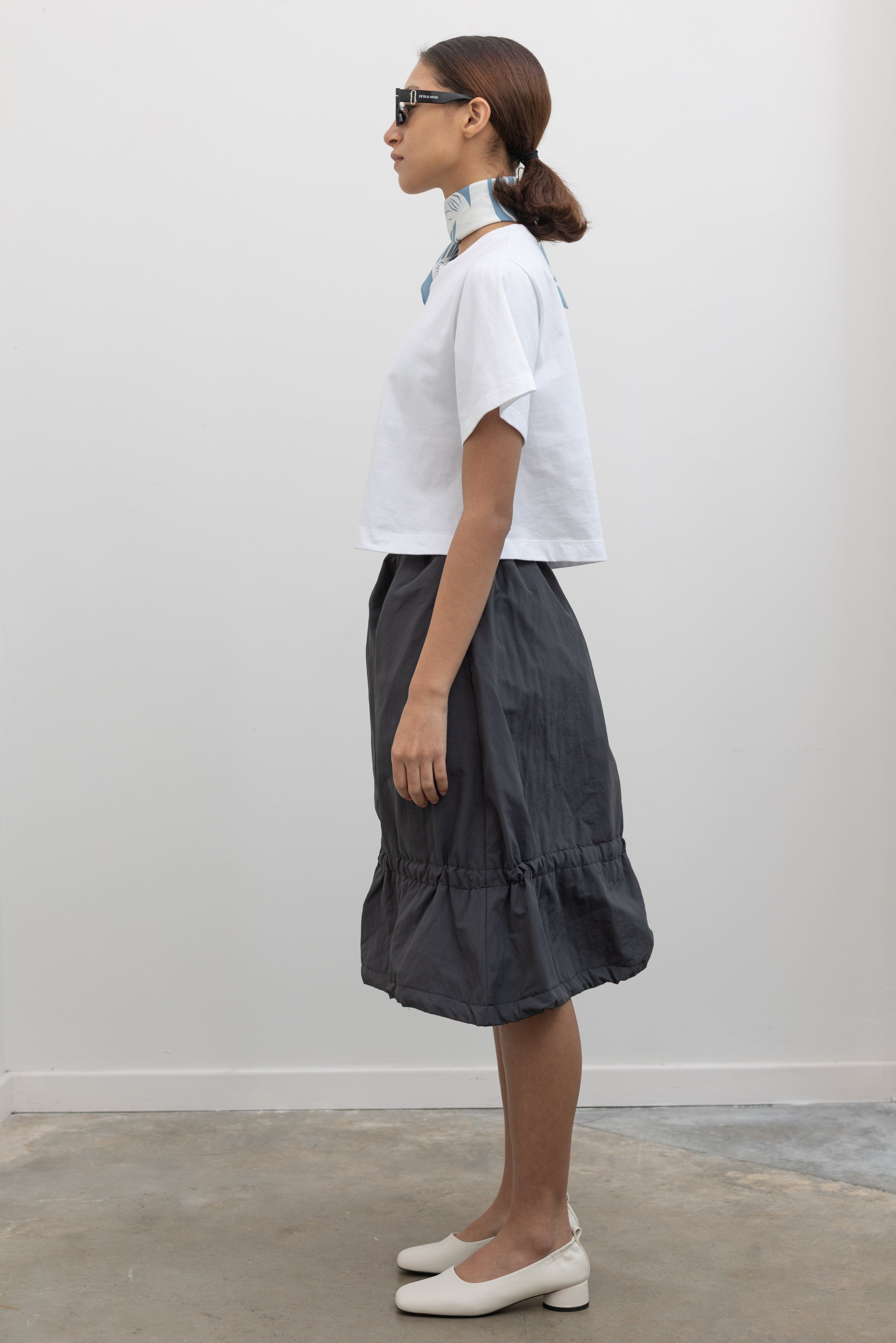 Full side view of a woman standing wearing Spring fashion in a Collective brand charcoal nylon bungee detail midi skirt with an Alas Eius label cropped white t-shirt at Alas Eius. 