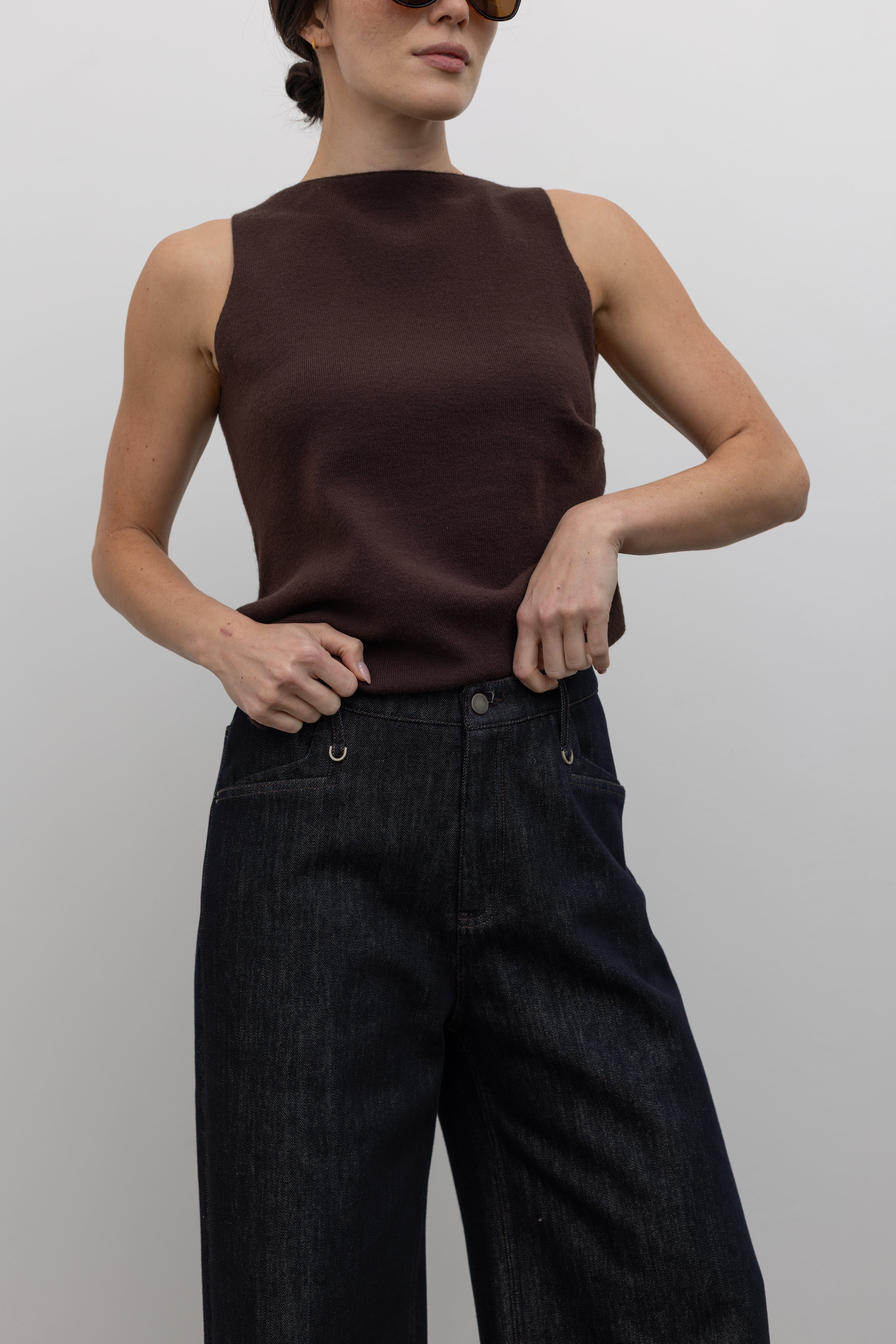 Close-up front view of a woman standing wearing modern 
minimalist fashion in a Collective sleeveless dark chocolate shirring knit top featuring a softly textured knit with subtle shirring and an asymmetrical hem paired with dark wash denim wide leg jeans at Alas Eius. 