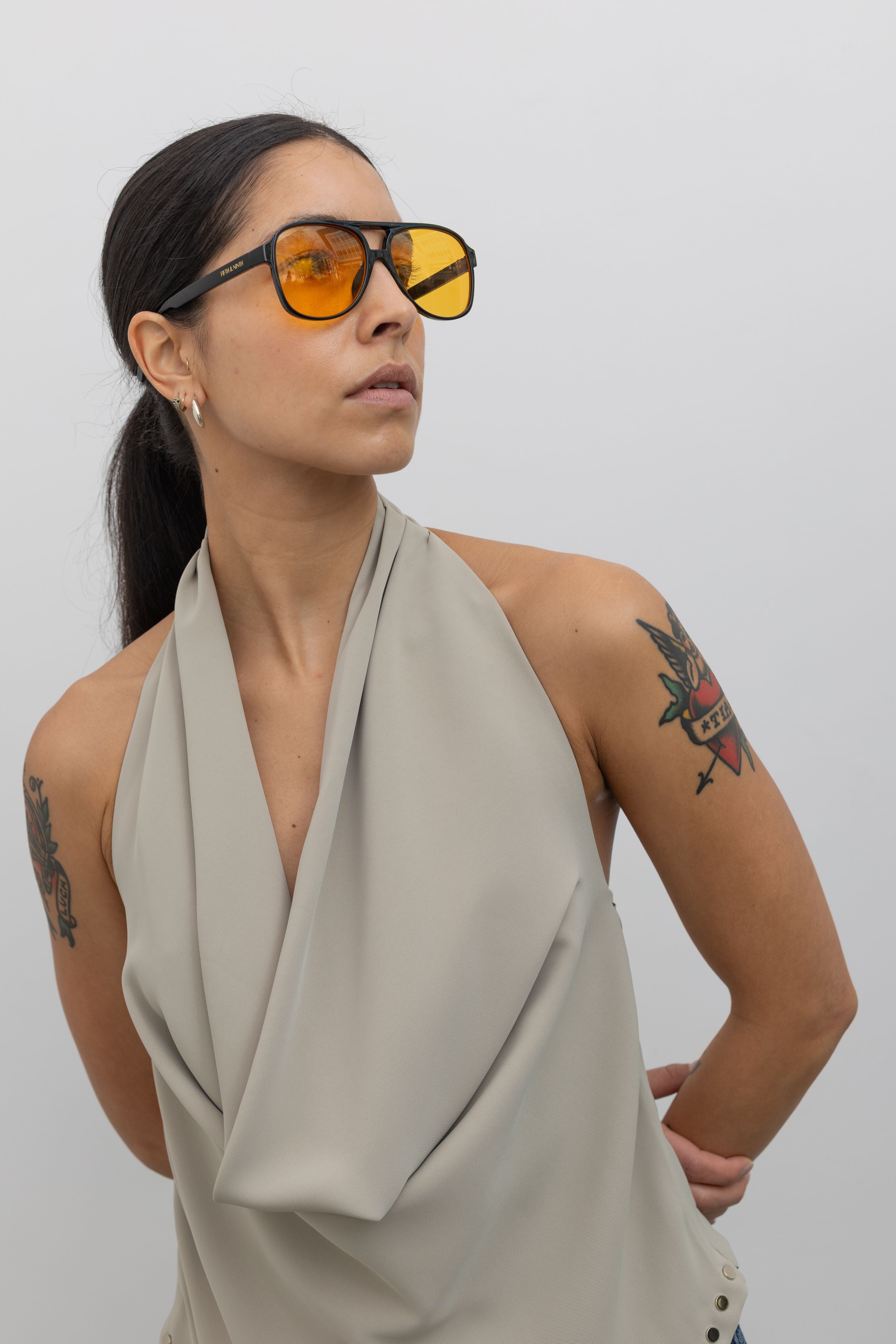 Close-up view of a woman wearing an edgy yet chic top in a Collective brand beige halter neck top designed with a low cowl drape, a v-cut hem that is lined with silver stud hardware, a side zipper closure, and exposed back at an Alas Eius Store.