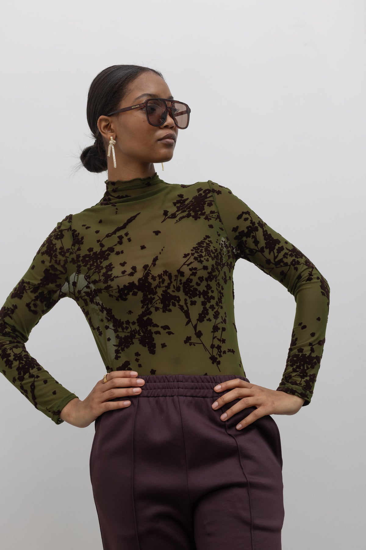 Woman posing with her hands on her hips wearing contemporary women's fashion in a Collective label olive green mesh high neck lettuce hem top featuring a raised floral printed throughout at an Alas Eius store. 