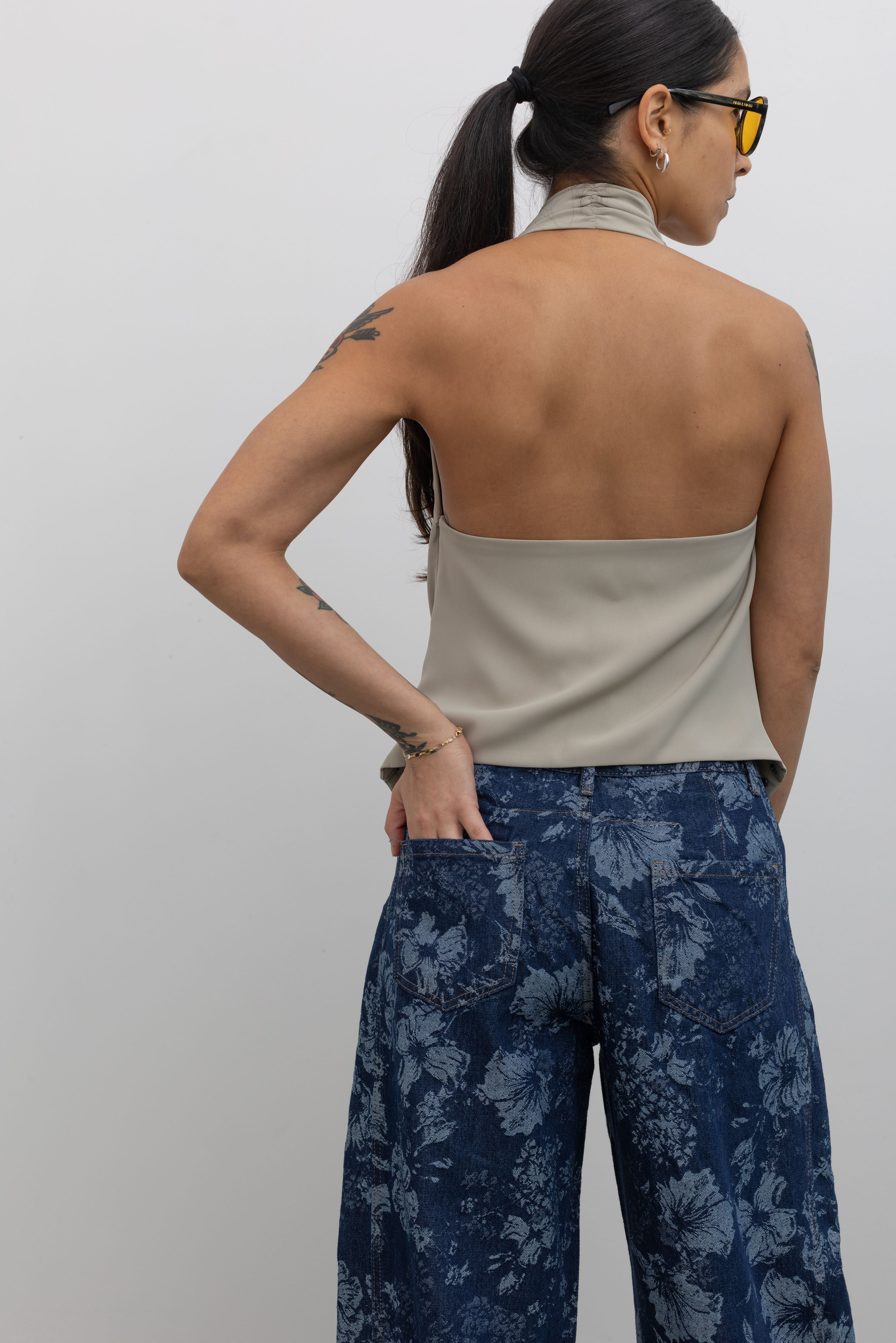 Back view of a woman wearing an edgy yet chic top in a Collective brand beige halter neck top designed with a low cowl drape, a v-cut hem that is lined with silver stud hardware, a side zipper closure, and exposed back at an Alas Eius Store.