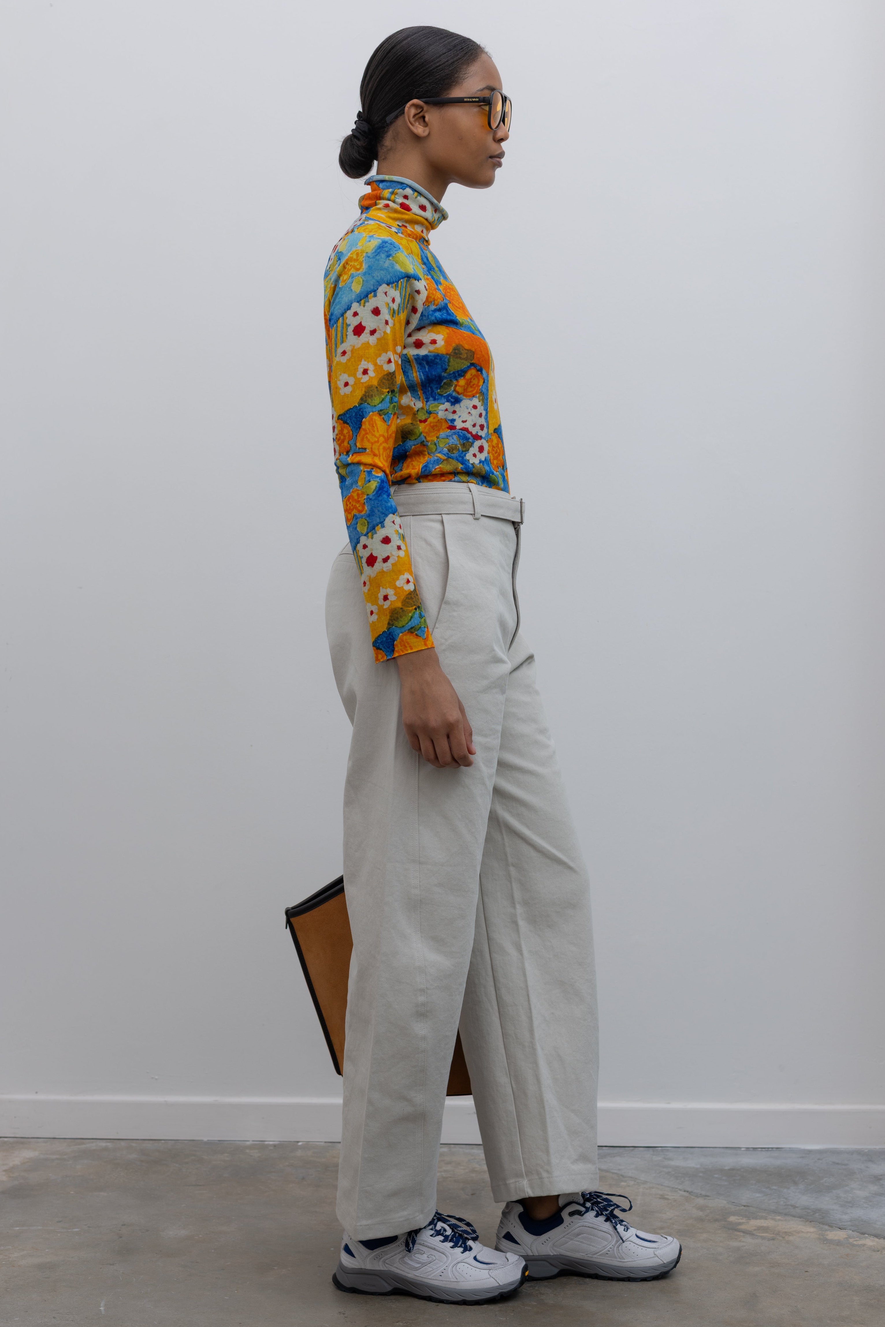 Side view of a woman wearing weekend fashion in a pair of Collective brand light gray curved leg belted chino pants with a vibrant orange and blue floral printed long sleeve top paired with white and blue sneakers at Alas Eius.