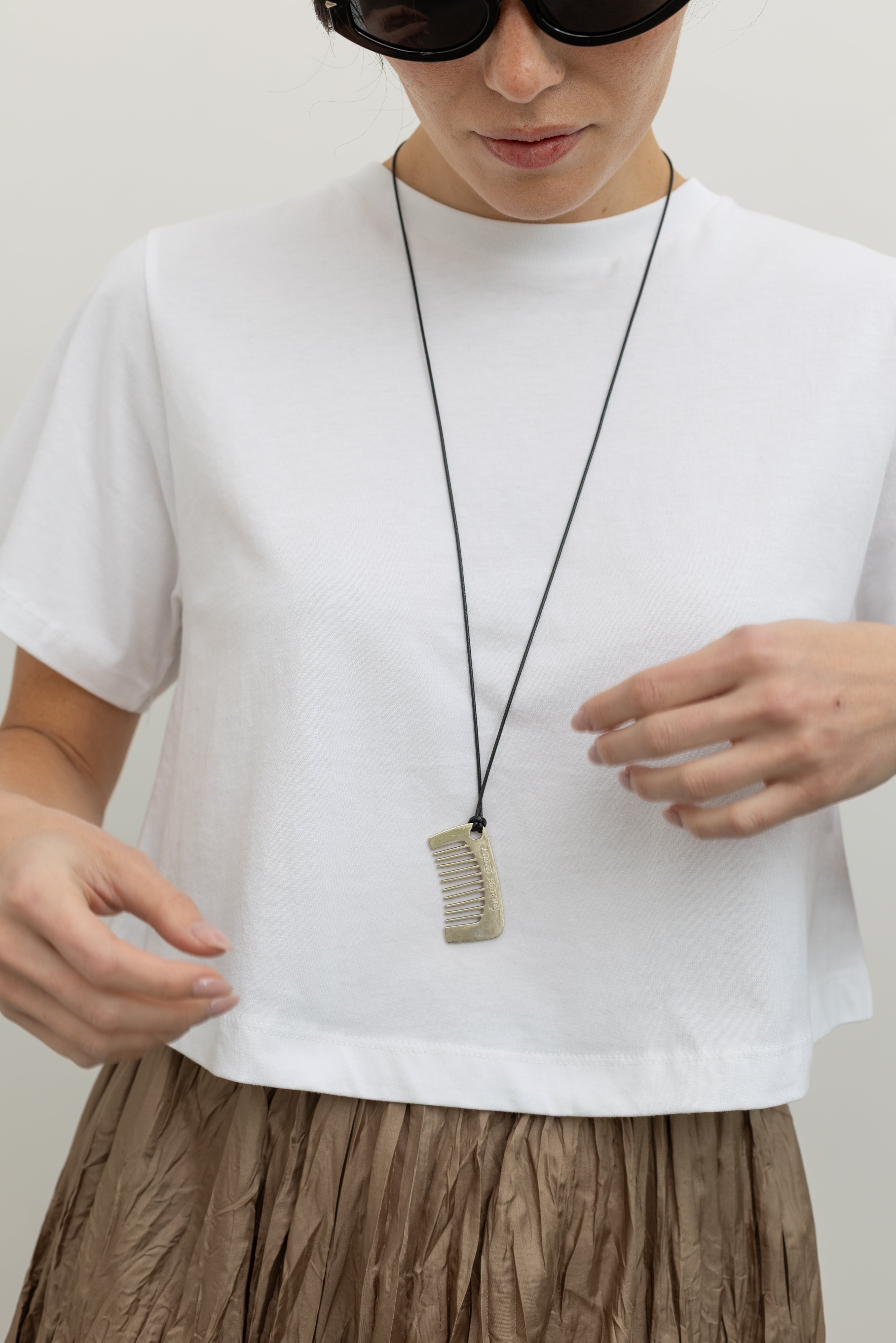 Woman wearing trendy Collective brand stainless steel silver comb pendant on an adjustable leather cord with “Atelier Edition” engraved on one side paired with an Alas Eius label white cotton cropped t-shirt, sunglasses, and crinkled maxi skirt at an Alas Eius store.