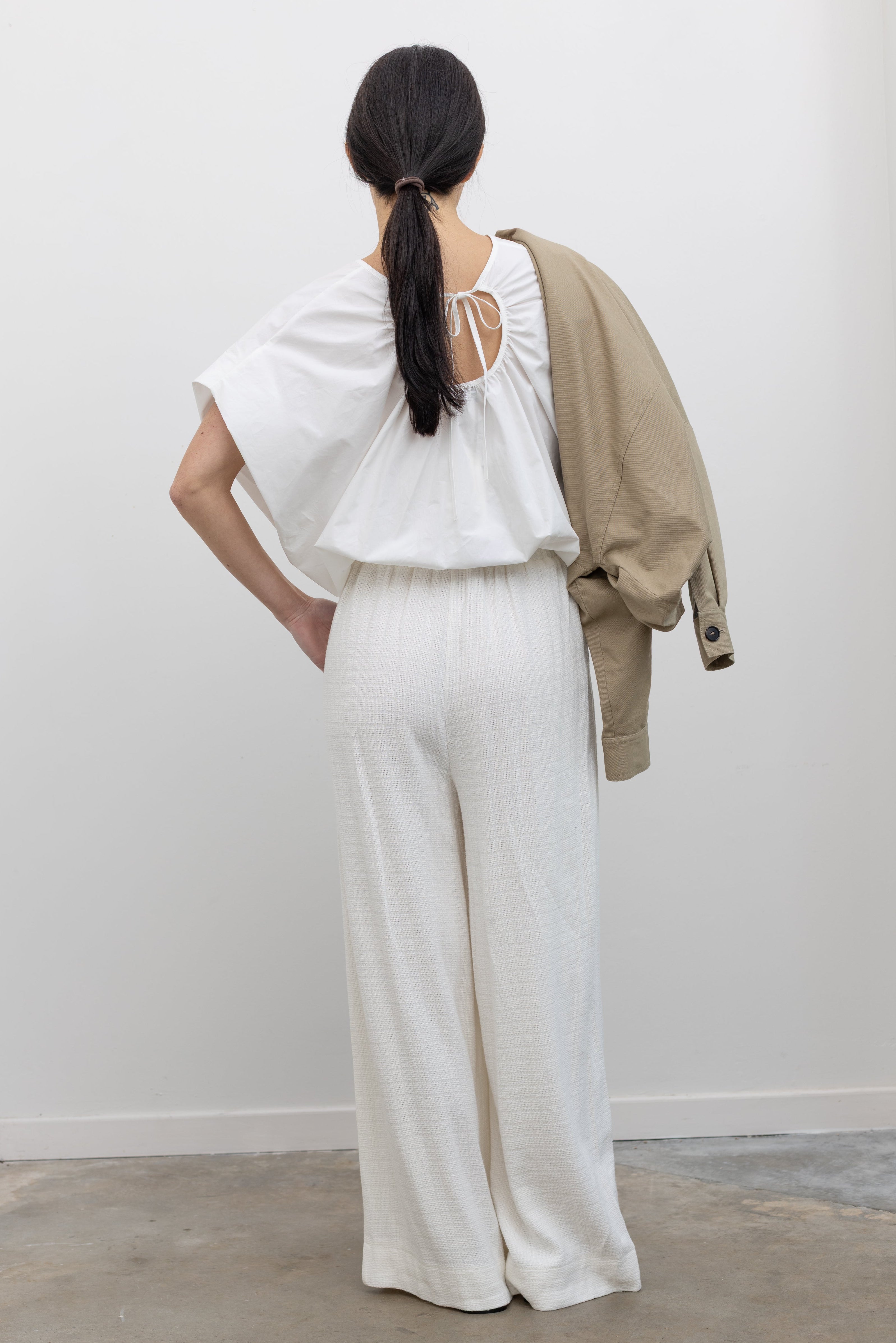 Full length back view of woman in motion wearing chic spring fashion in Collective Ivory Linen Wide Leg Pants featuring an elastic waistband and side seam pockets paired with an oversized short sleeve top and strappy leather sandals at Alas Eius.