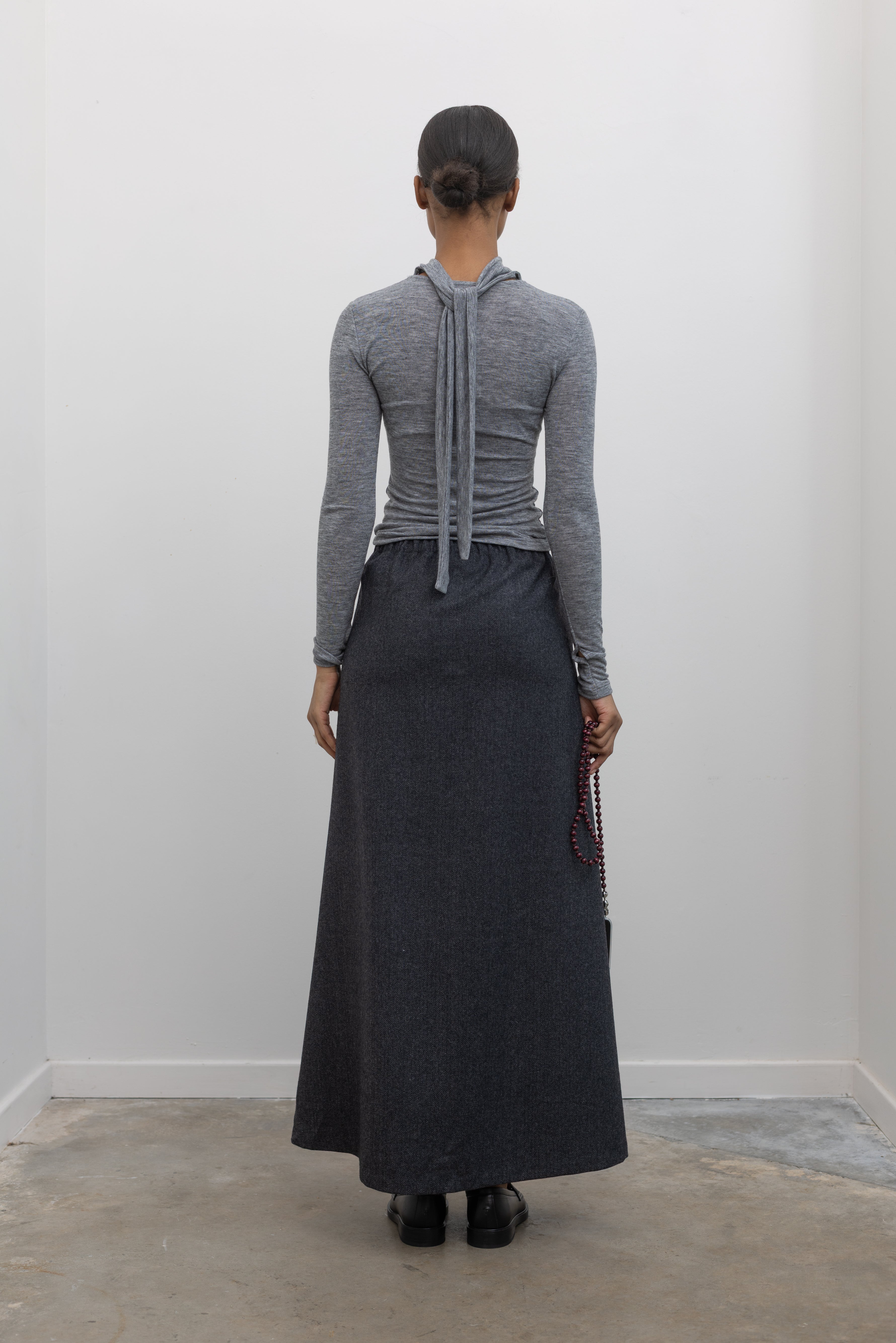 Full length back view of a woman wearing a sophisticated and refined outfit featuring an Collective label charcoal gray herringbone weave A-line maxi skirt paired with a fitted heather gray mock neck top at Alas Eius. 
