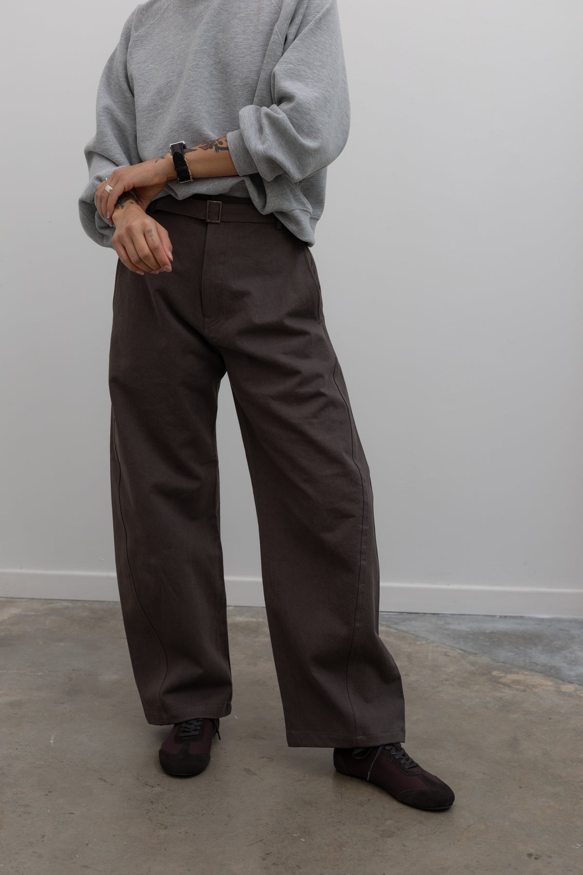 Close-up front view of a woman wearing weekend fashion in a pair of Collective brand brown curved leg belted chino pants with a casual heather gray sweatshirt and brown suede ballet inspired sneakers at an Alas Eius store. 