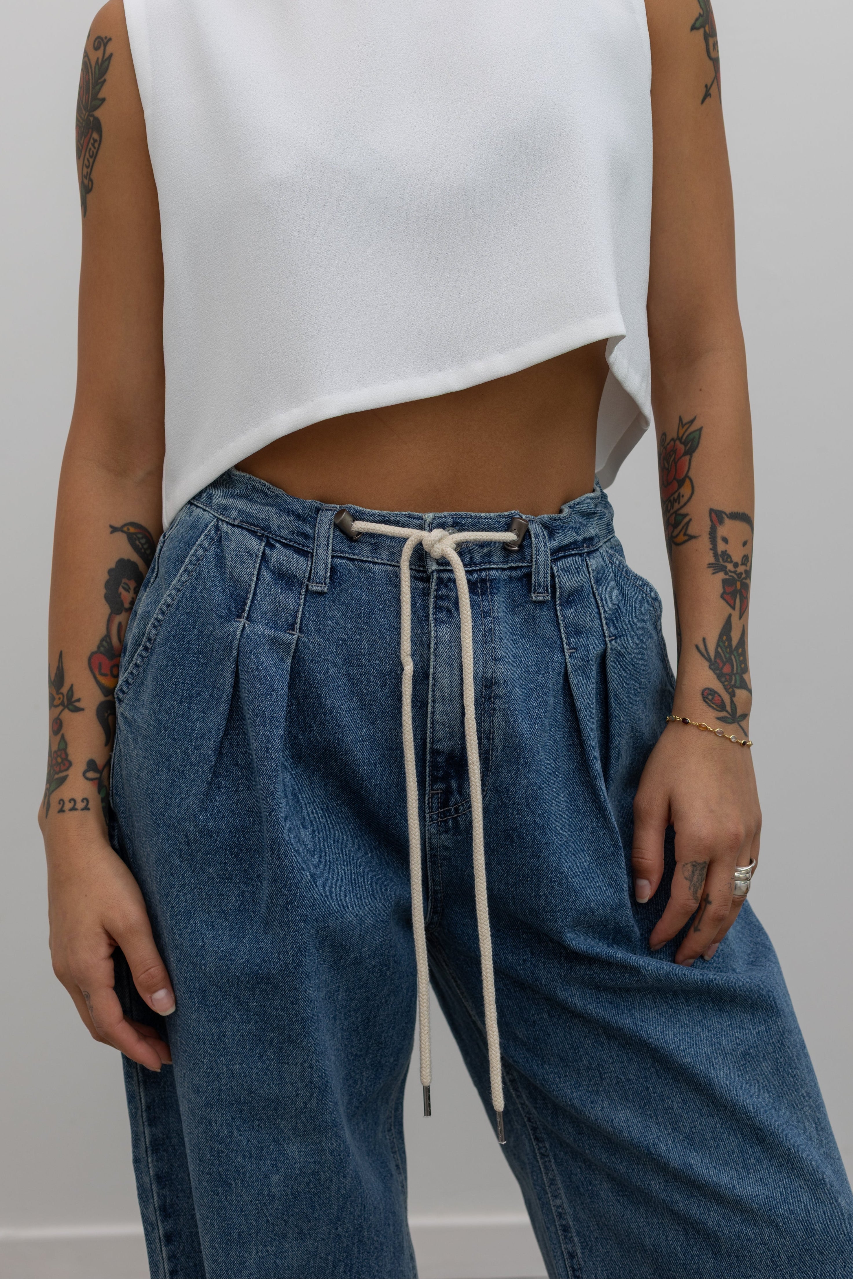 Woman wearing pleated front jeans featuring a drawstring waist and a mid-rise silhouette paired with a white cropped top at an Alas Eius store. 