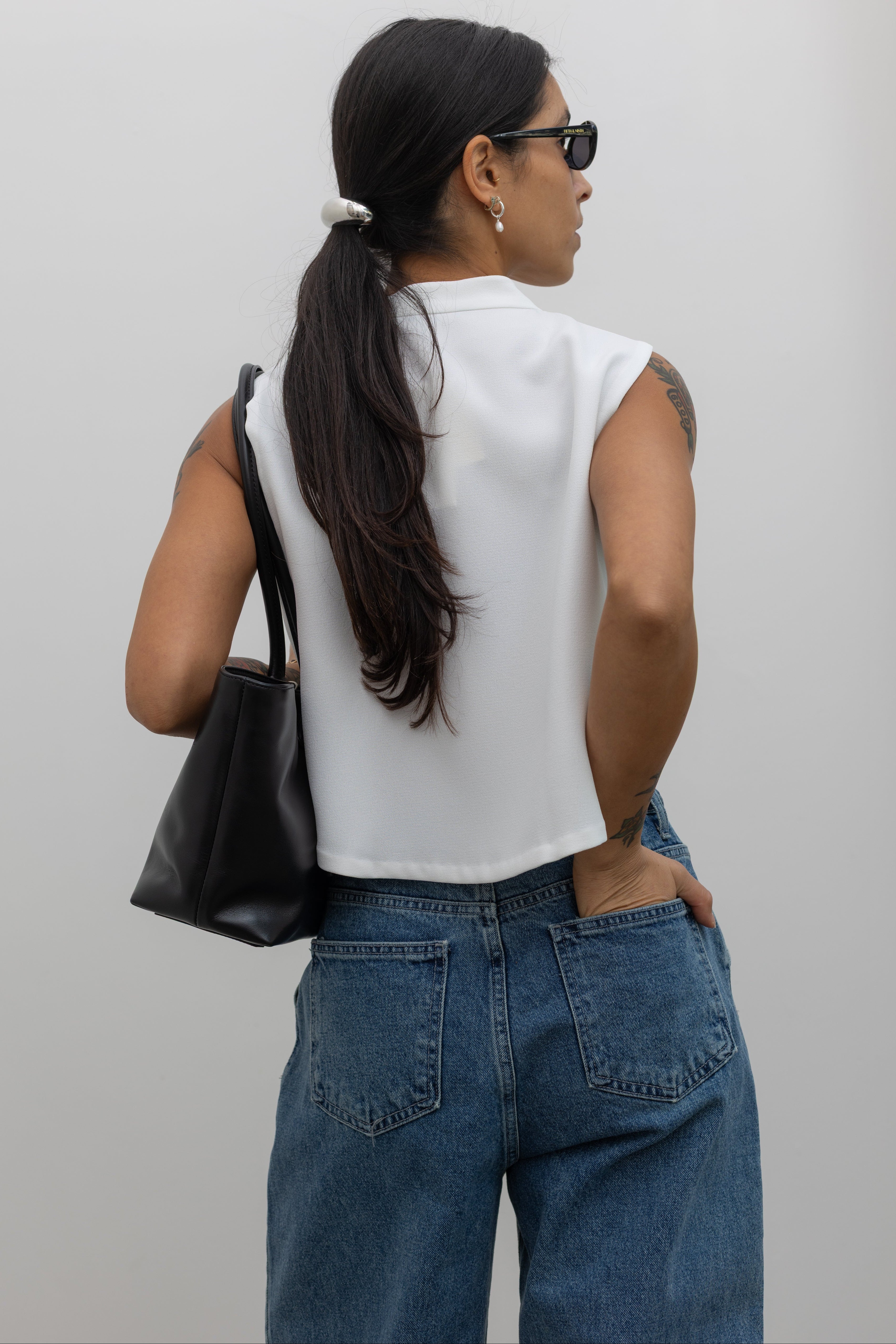 Back view of a woman wearing flattering drawstring waist, mid-rise light wash denim jeans with a white copped sleeveless top and a black leather bag Alas Eius. 
