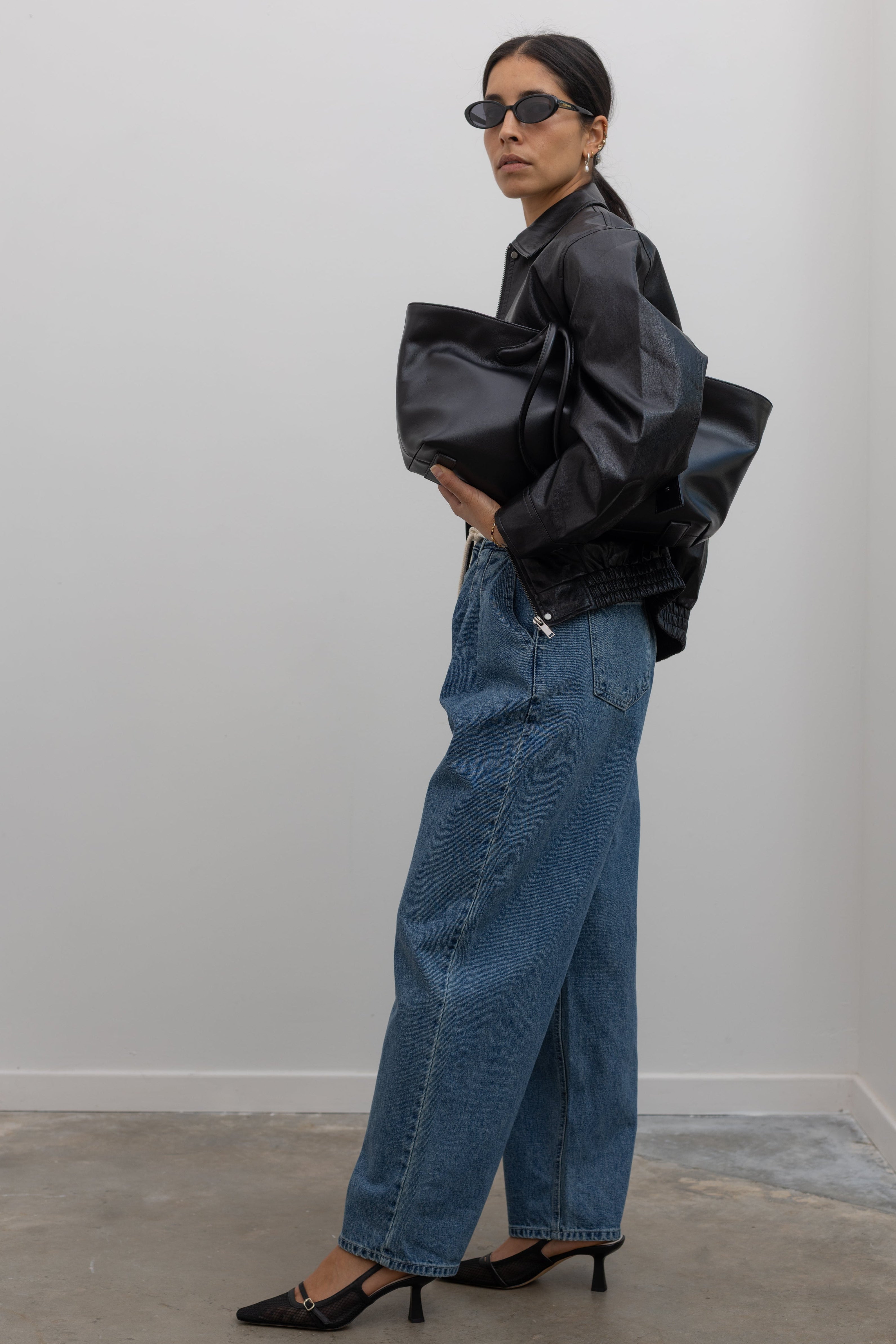 Side view of a woman wearing pleated front jeans featuring a drawstring waist, a mid-rise silhouette, and a straight leg cut paired with slingback heels and a black leather jacket at Alas Eius. 