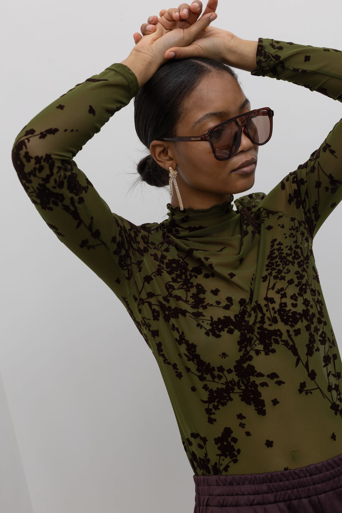 Woman posing with her hands over her head wearing the perfect date night top in a Collective label olive green mesh high neck lettuce hem top featuring a raised floral printed throughout at an Alas Eius store. 