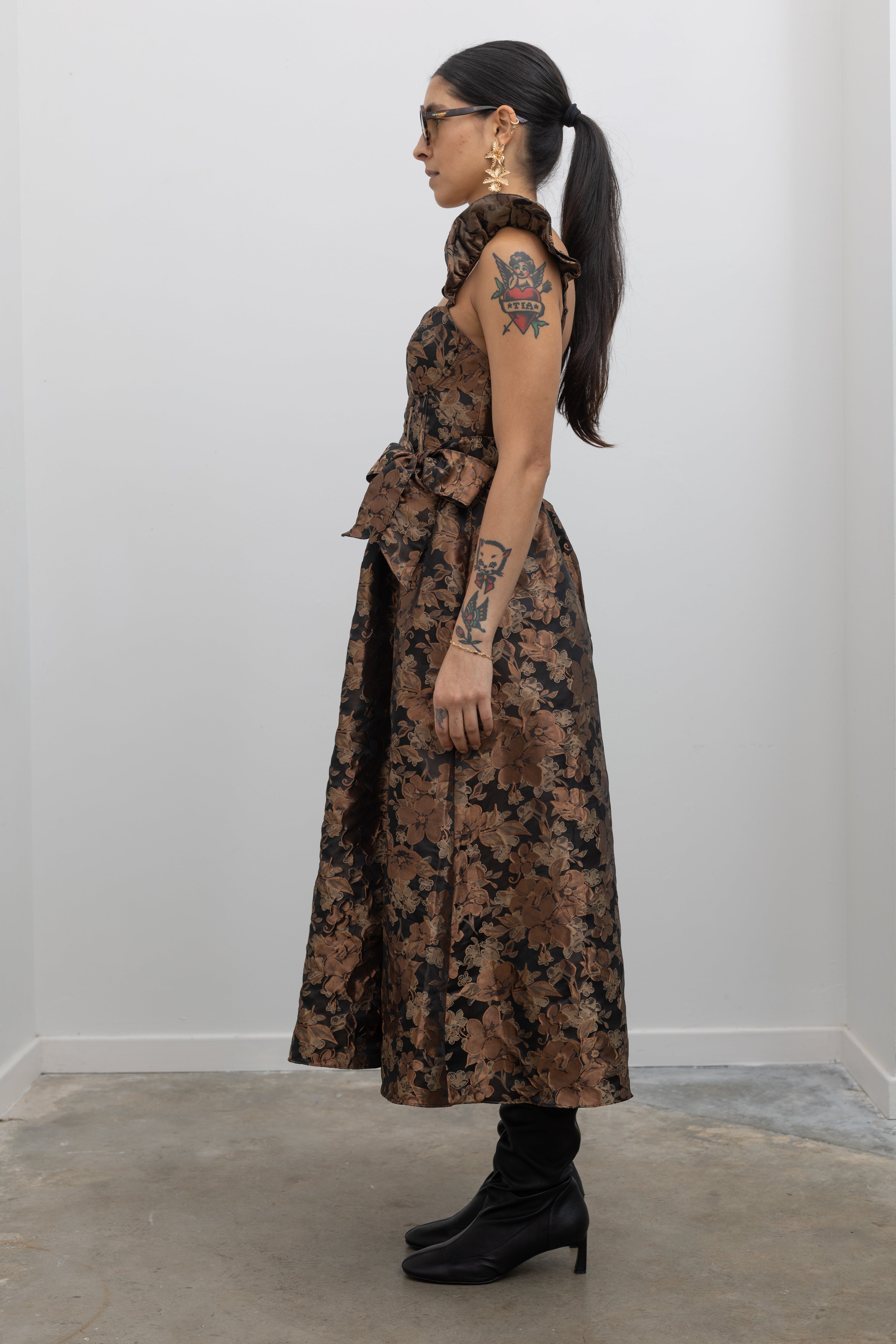 Full side view of a woman wearing flattering feminine Collective label bronze floral jacquard corset midi dress with slouchy black leather heeled boots at Alas Eius. 