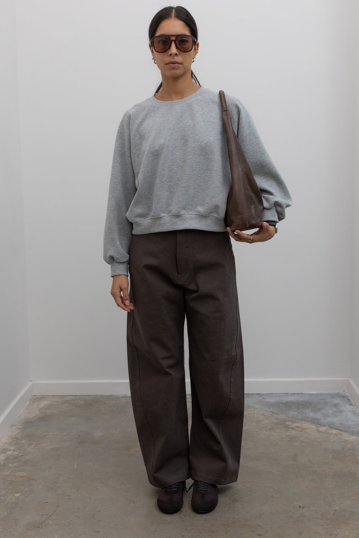 Front view of a woman wearing weekend fashion in a pair of Collective brand brown curved leg belted chino pants with a casual heather gray sweatshirt and brown suede ballet inspired sneakers at an Alas Eius store. 
