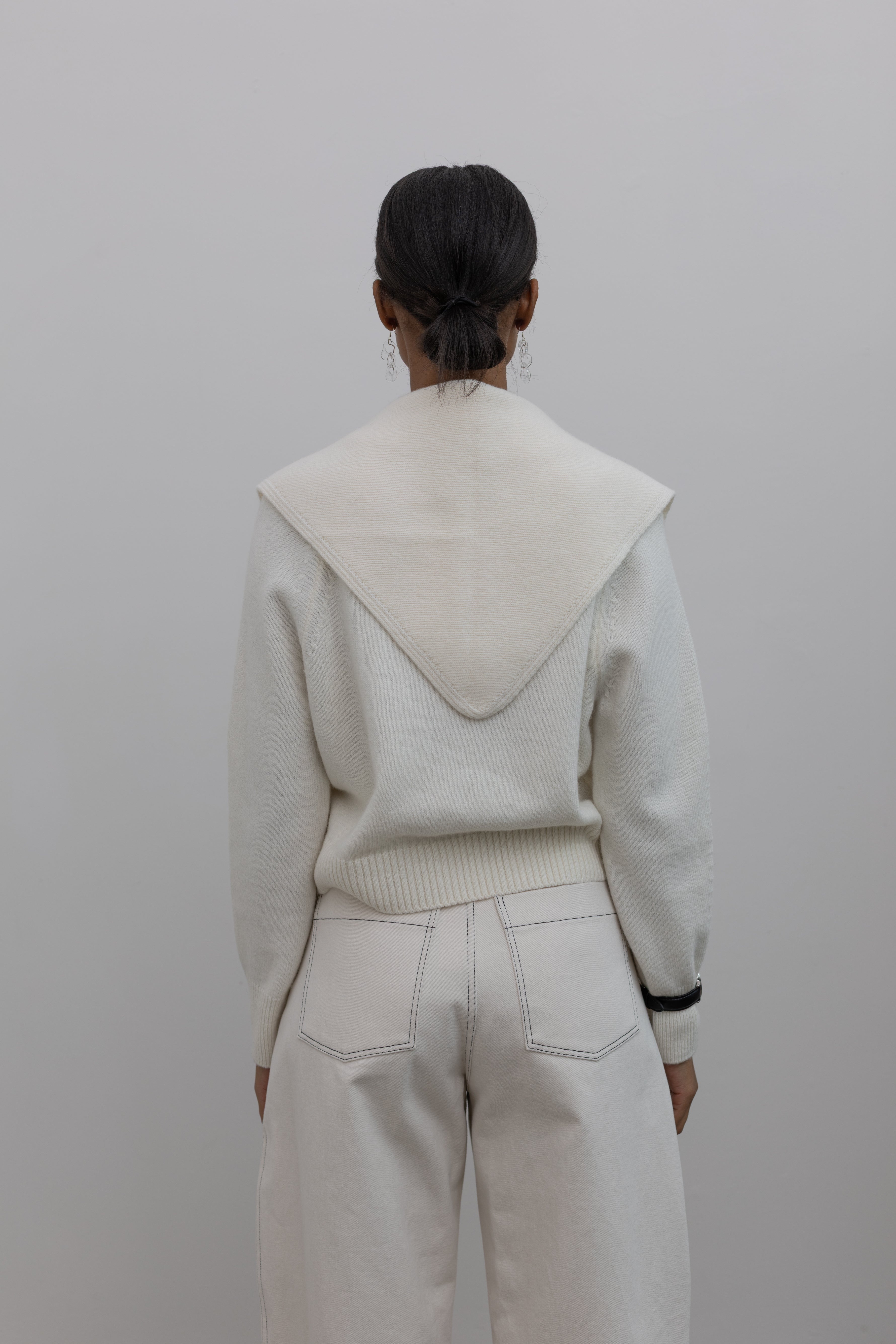 Partial back view of a woman standing wearing an everyday capsule wardrobe outfit in a white long sleeve top with Collective label cream curved barrel leg pants featuring black contrast stitching paired with a black leather belt with silver hardware and black leather loafers at Alas Eius. 