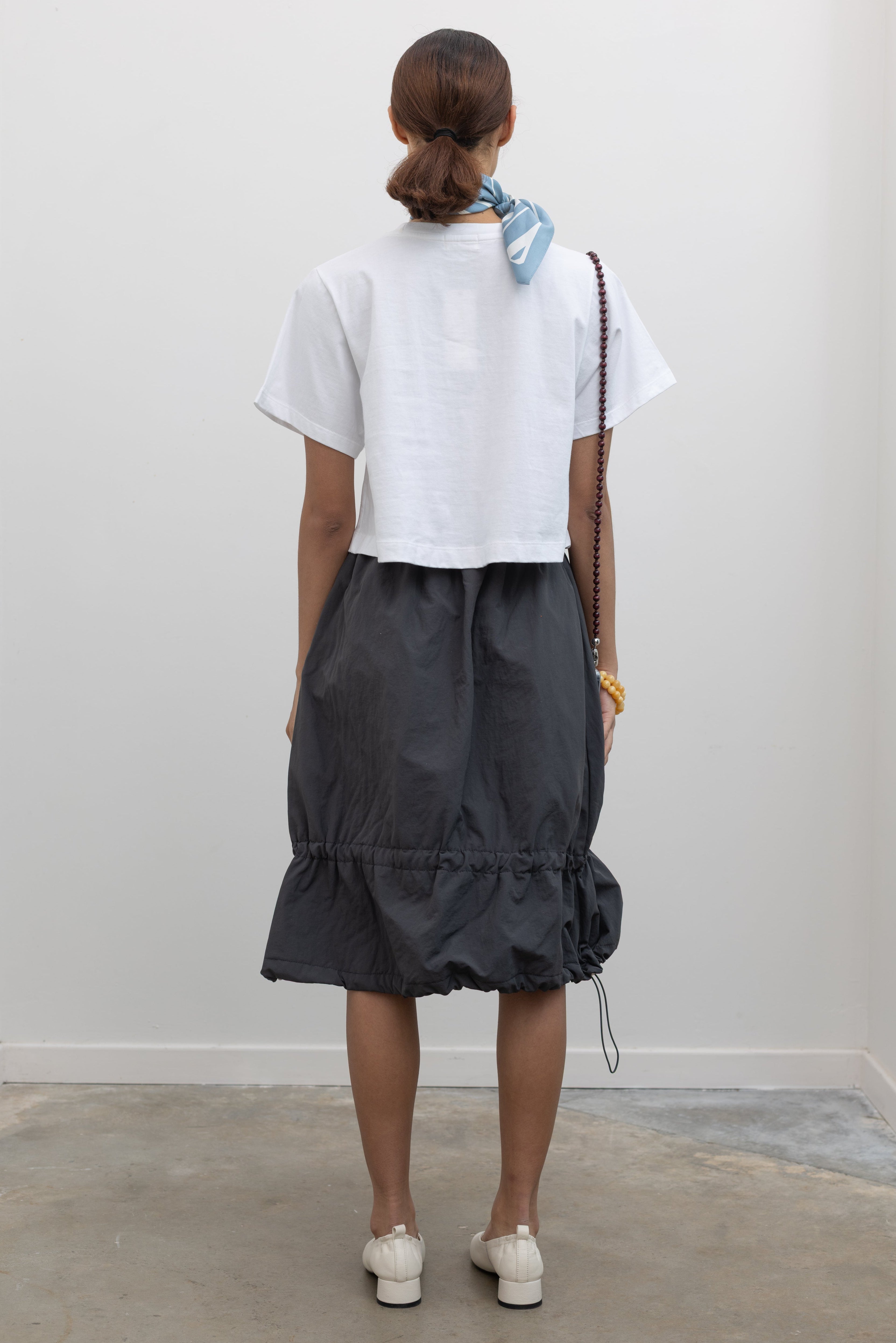 Full back view of a woman standing wearing Spring fashion in a Collective brand charcoal nylon bungee detail midi skirt with an Alas Eius label cropped white t-shirt at Alas Eius. 