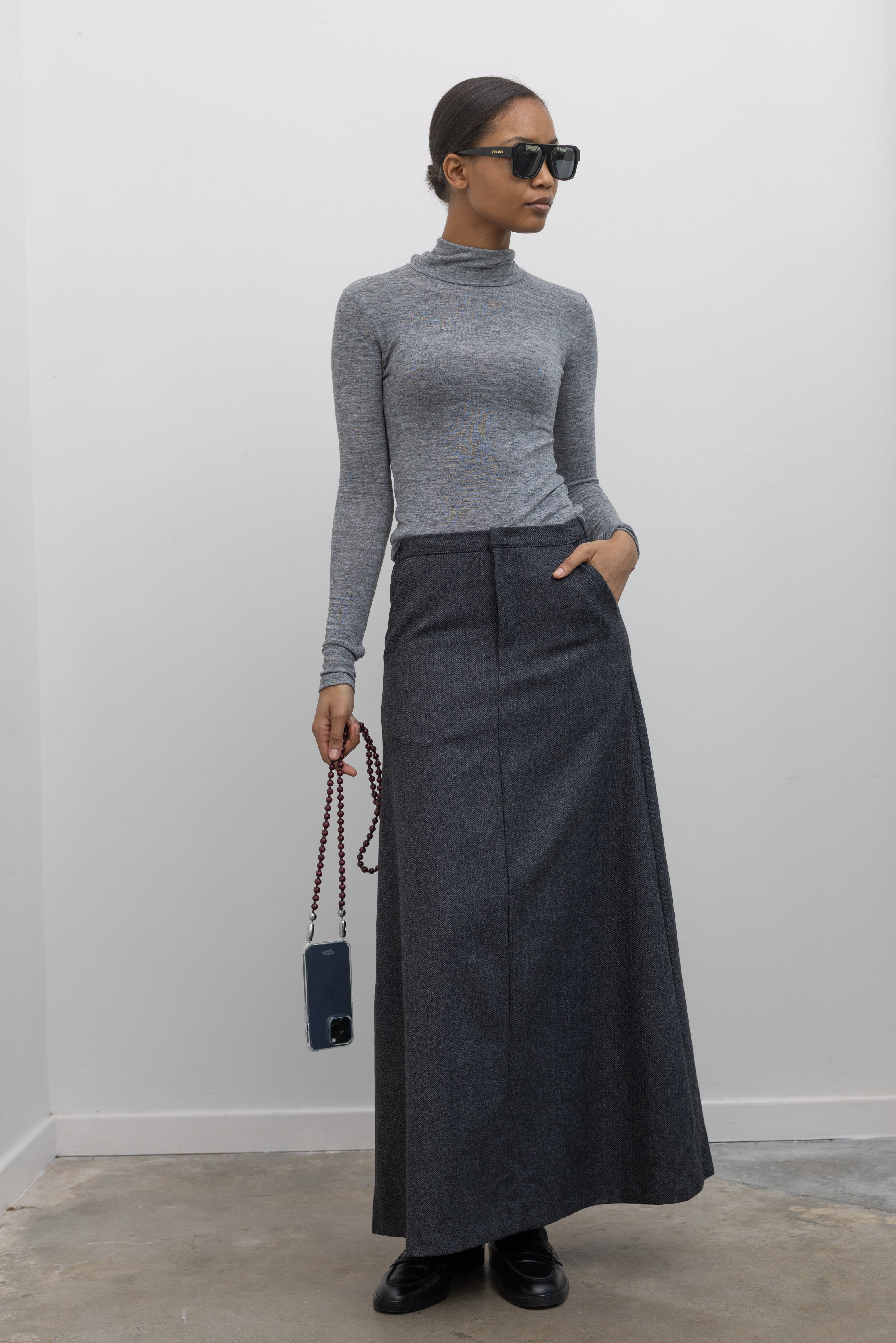 Full length view of a woman wearing a sophisticated and refined outfit featuring an Collective label charcoal gray herringbone weave A-line maxi skirt paired with a fitted heather gray mock neck top at Alas Eius. 