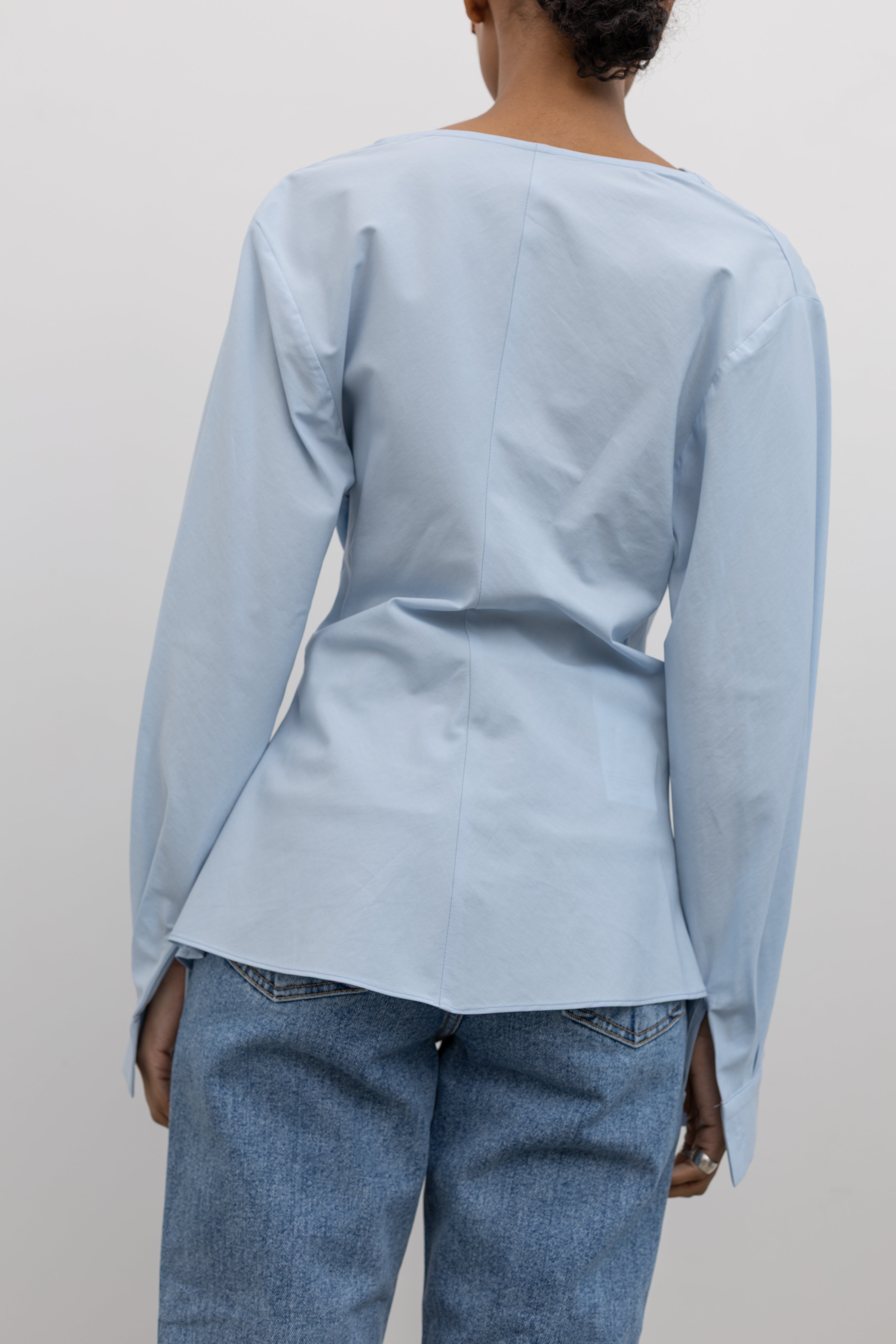 Back view of a Woman wearing chic and architectural spring fashion in a Collective label sky blue cotton long sleeve top featuring a fitted natural waistline with a side draped motif creating an asymmetrical hem and cowl neckline paired with light wash denim jeans at Alas Eius. 