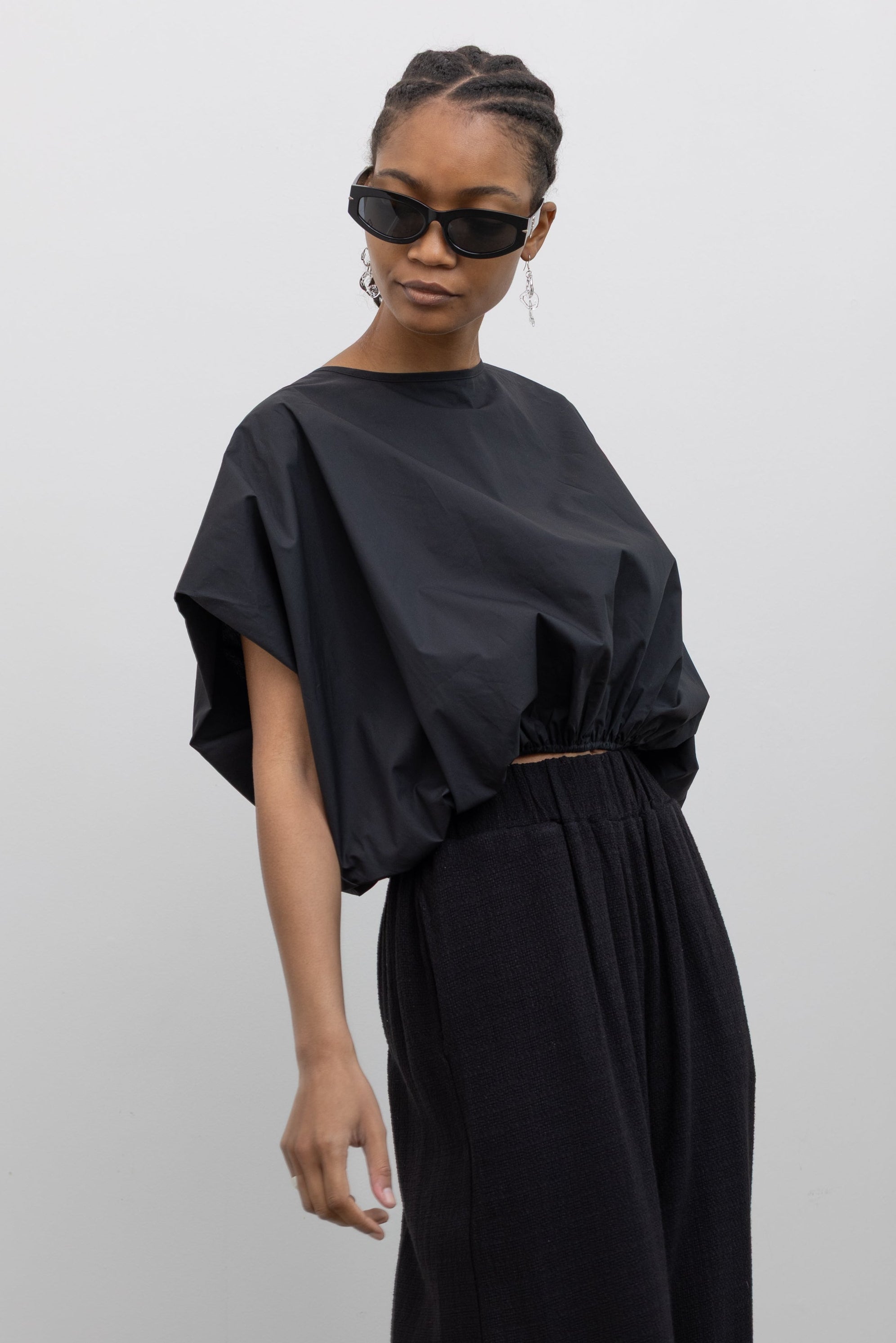 Close-up 3/4 view of a woman wearing Spring fashion in a sculptural  Collective Black Cotton billow cinched top featuring draped rounded sleeves, an elasticized waistband for a fitted torso and ribbon tie at the back neck at Alas Eius. 